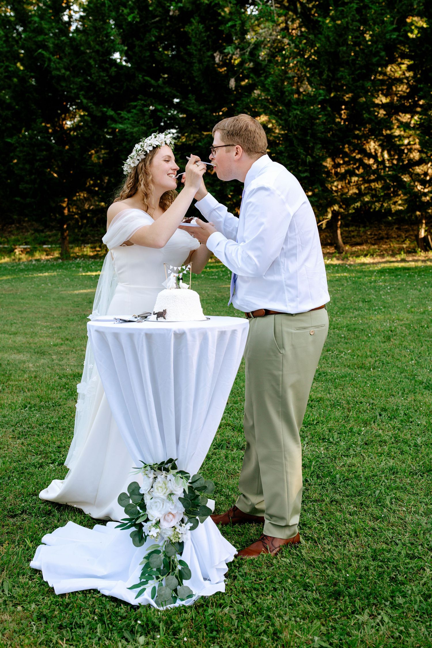 Bride and groom feed each other wedding cake at an outdoor reception on a sunny green lawn.