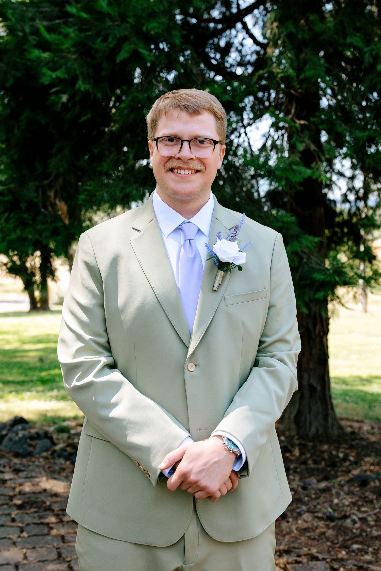 Smiling groom in a sage green suit with lavender tie and white boutonniere posing outdoors near lush green trees.