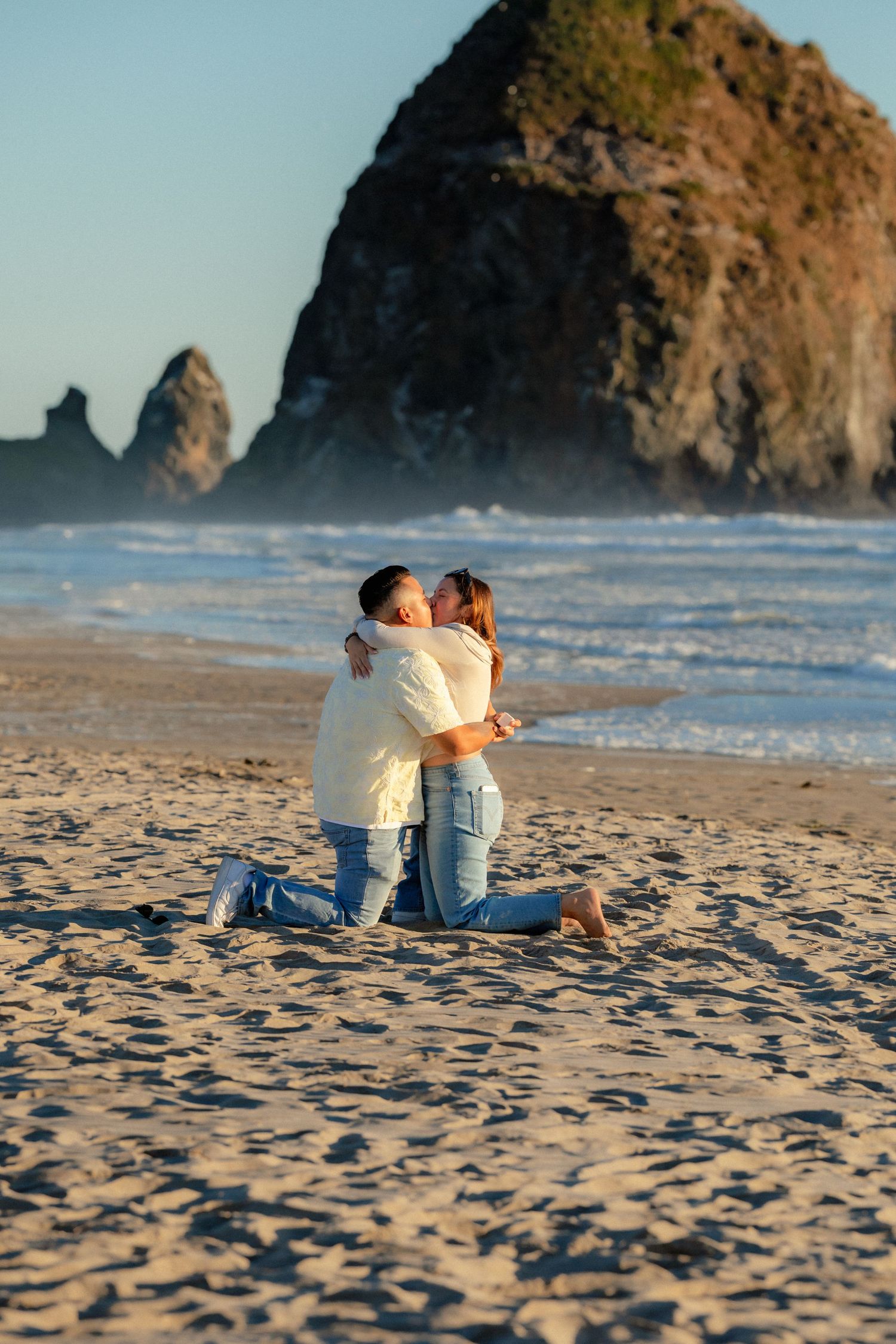 Couple kneeling and kissing on sandy beach with iconic Haystack Rock at Cannon Beach, Oregon in background.
