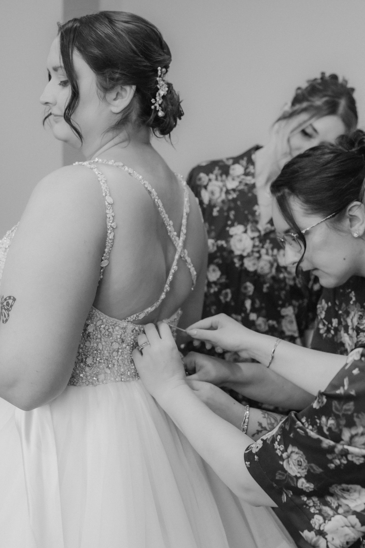 Black and white photo of bride getting wedding dress fastened by two women in floral dresses before ceremony.