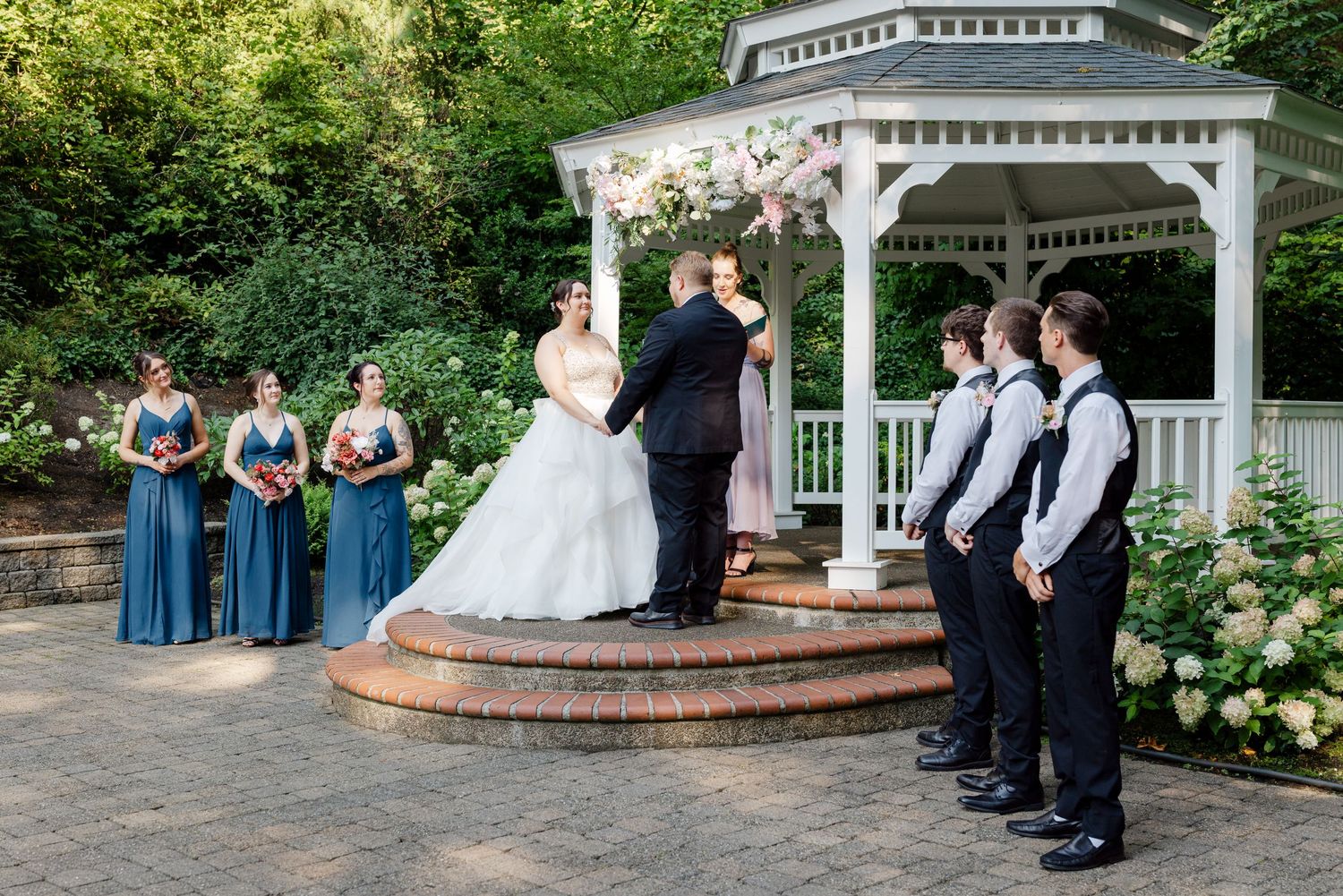 Bride and groom exchange vows at gazebo flanked by bridesmaids in blue dresses and groomsmen in black attire.
