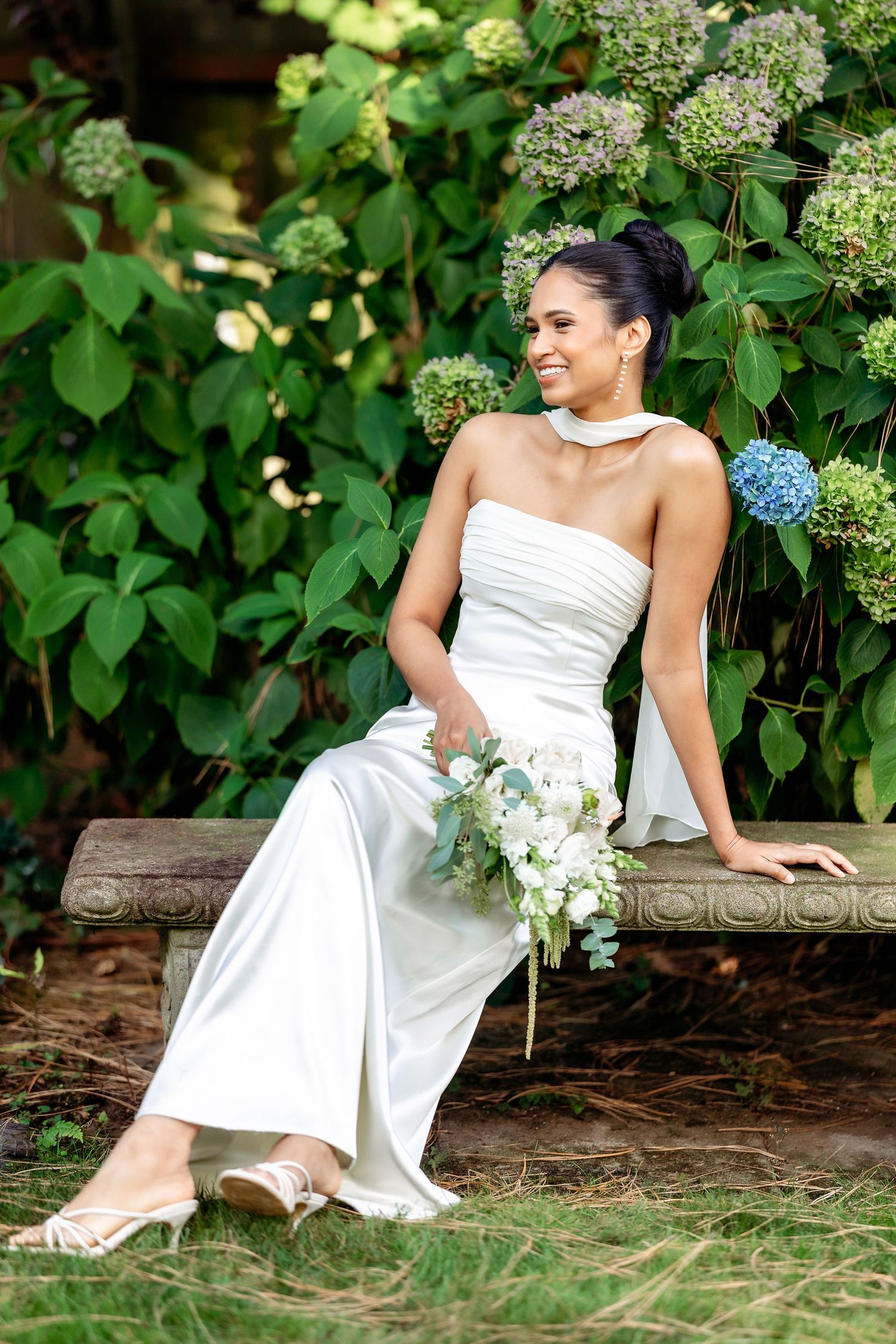 Bride in white dress posing on rustic bench surrounded by greenery.