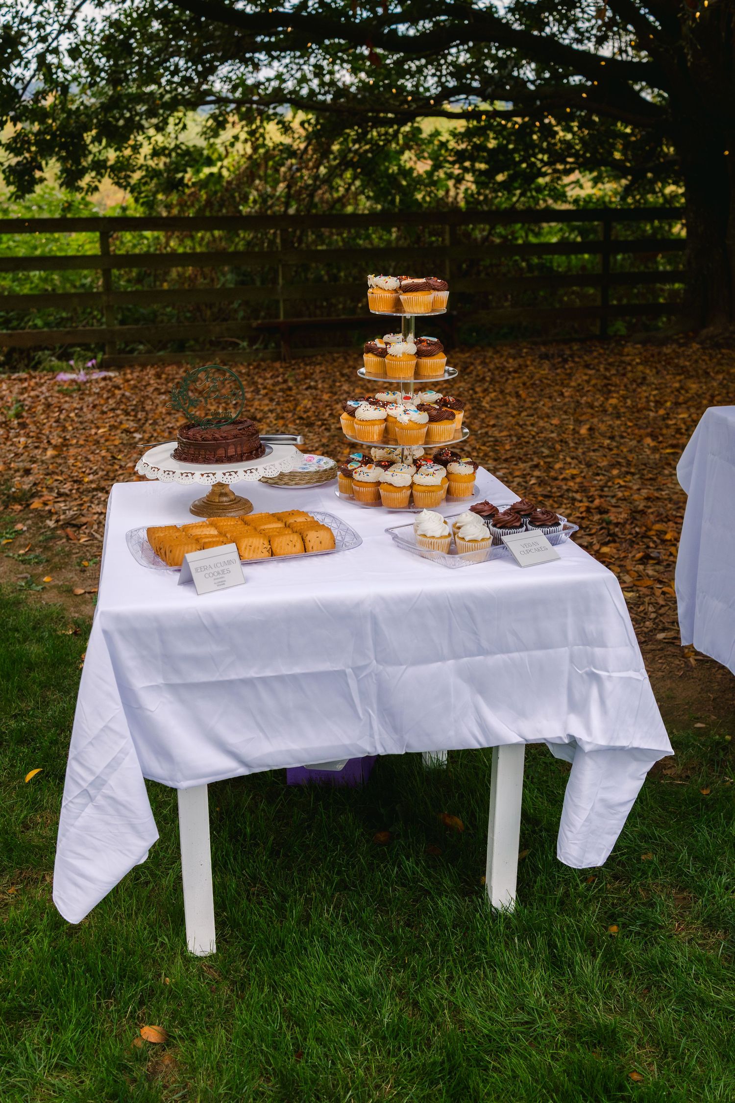 White tables covered in tablecloths display desserts and cupcakes arranged on tiered stands in an outdoor garden setting.