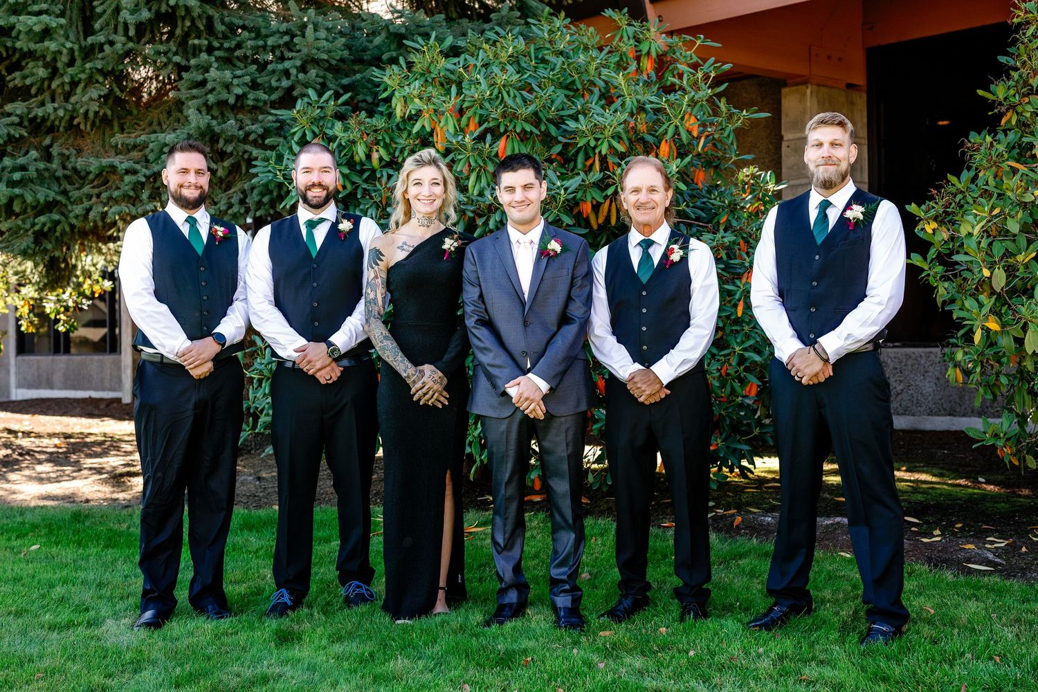 A row of groomsmen in black suits and vests pose together outdoors in front of greenery and trees.