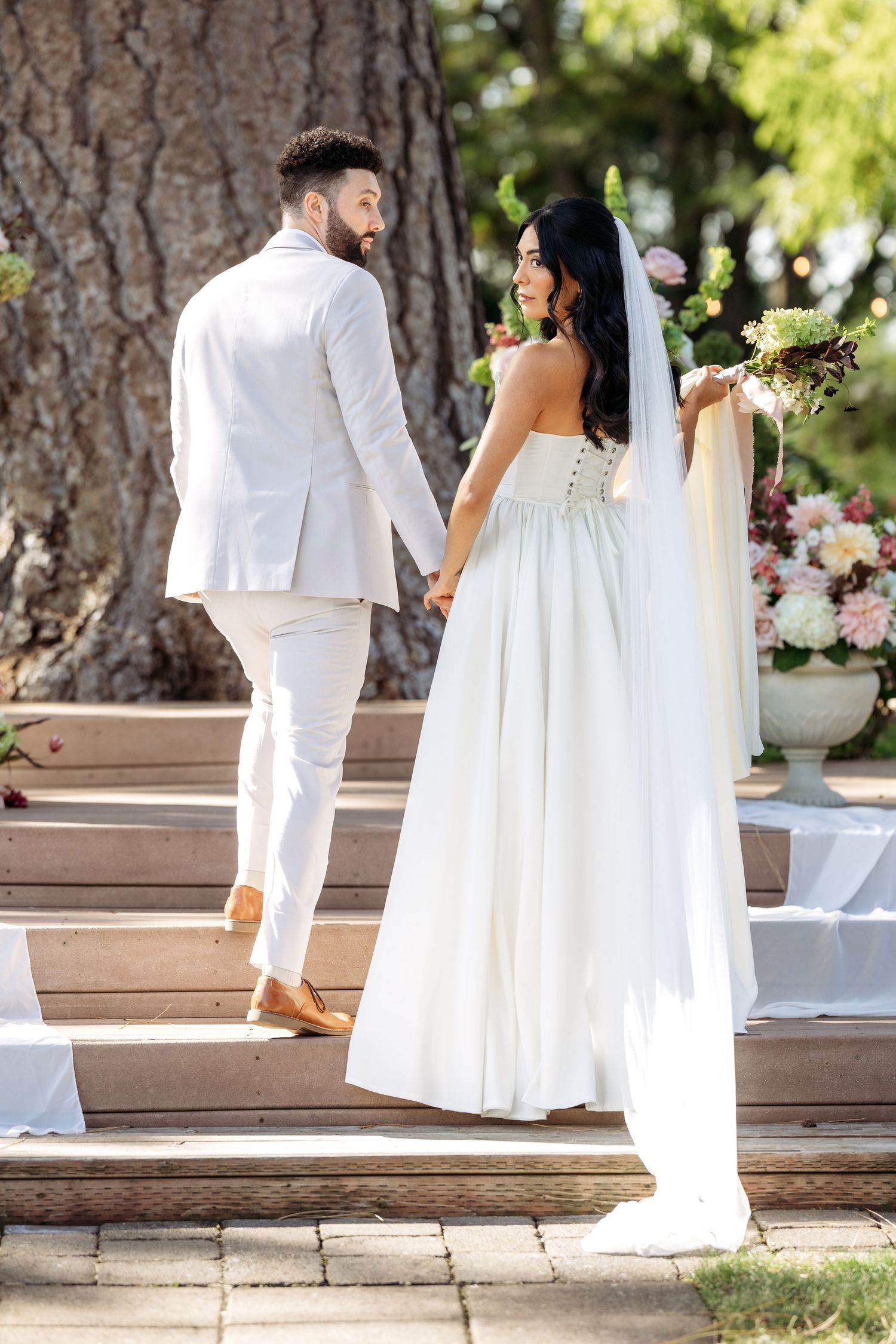 Romantic wedding couple in white attire poses together on outdoor steps in a garden setting.