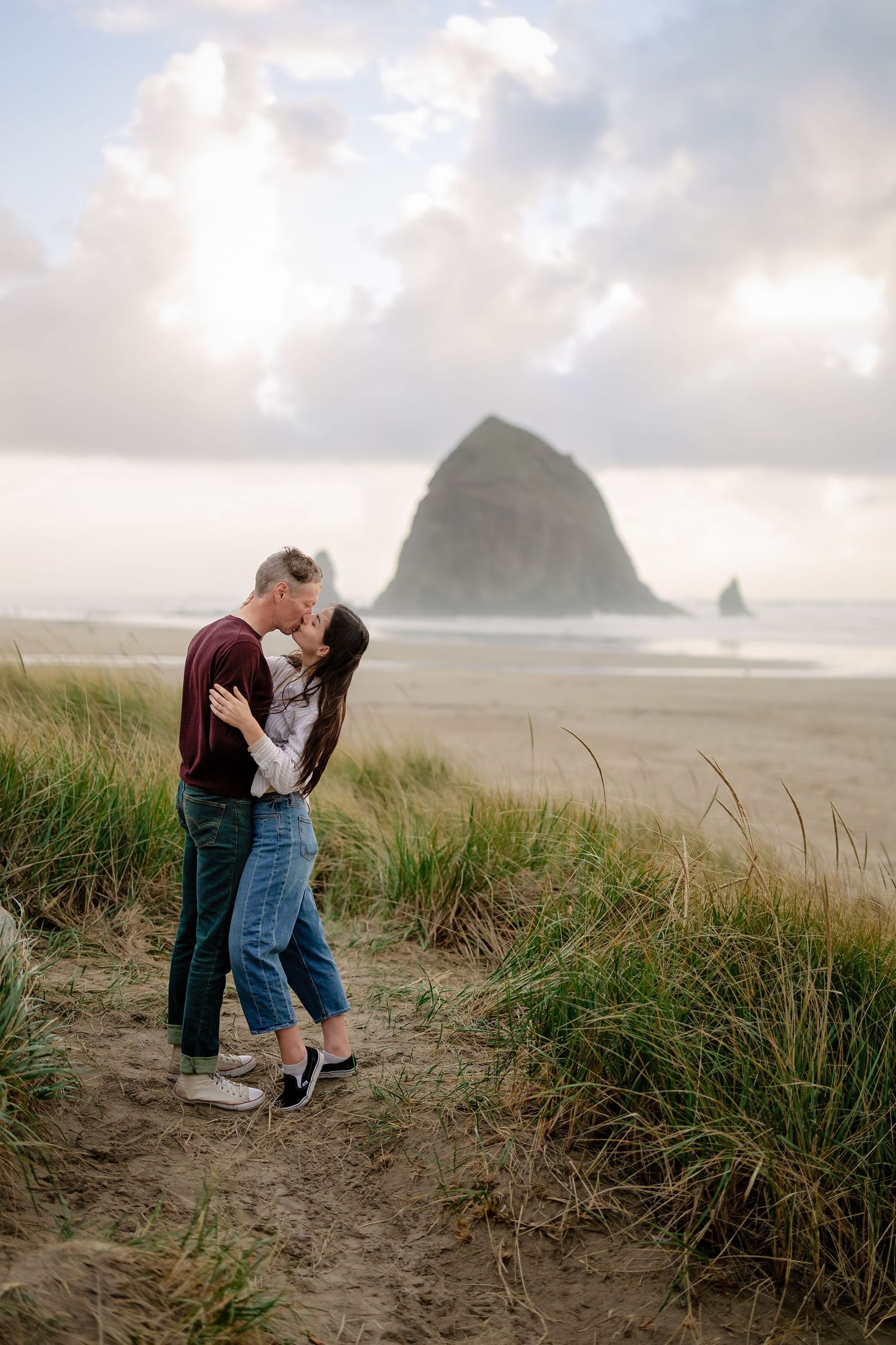 A couple shares an intimate moment embracing on a misty beach with tall grass and coastal rock formations in the background.
