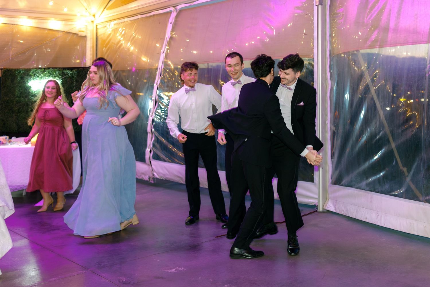 Wedding guests dancing and socializing on a purple-lit dance floor under a clear tent during an evening reception.
