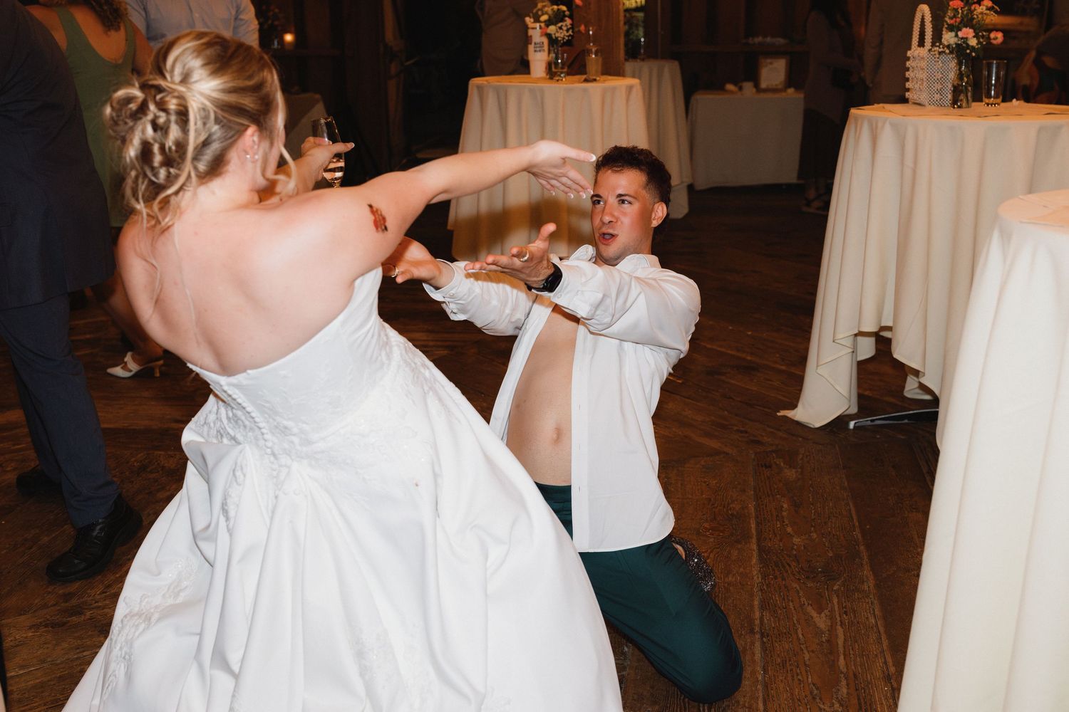 A bride in a white gown dances with a kneeling guest at a festive wedding reception.