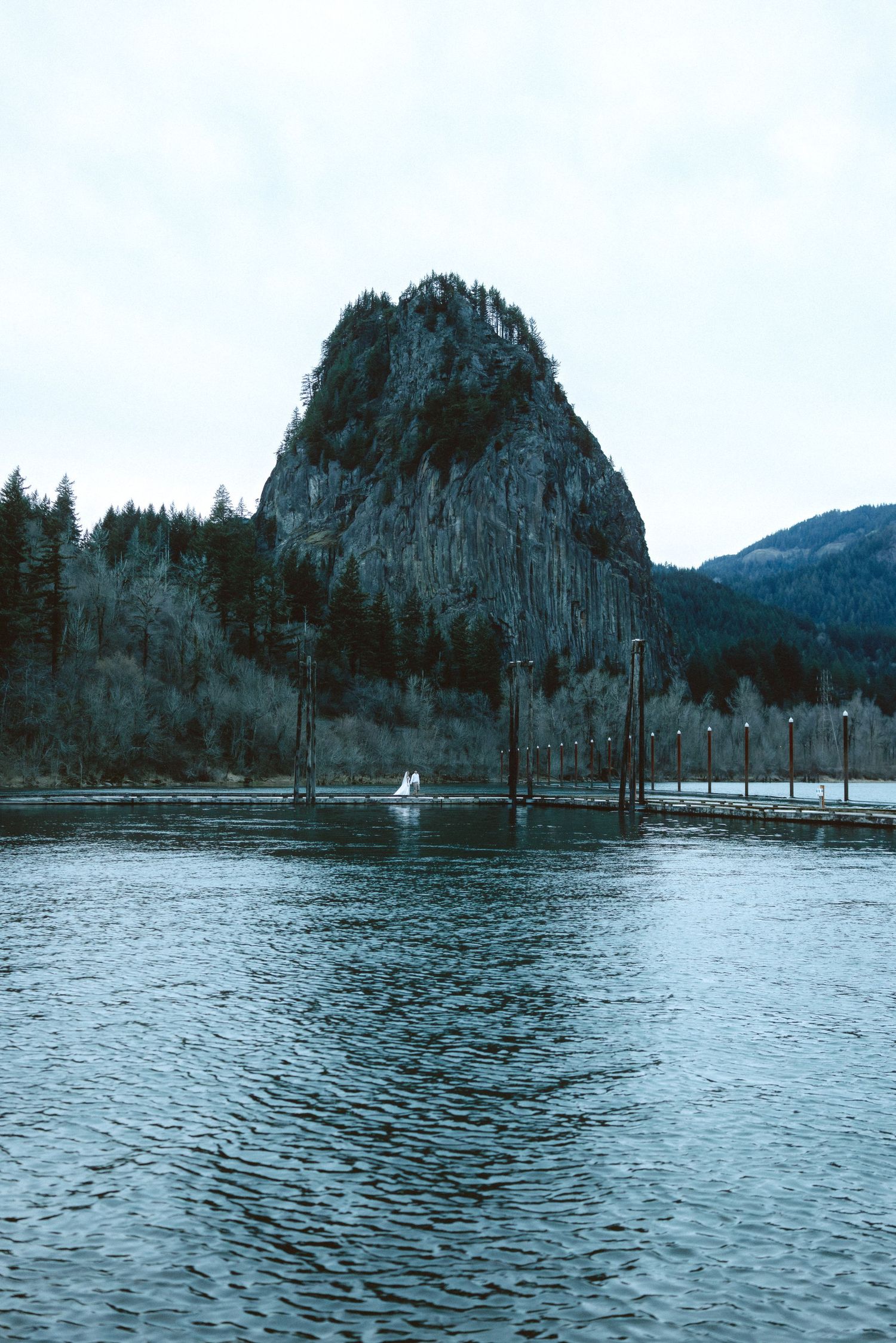 A panoramic view of a serene lake surrounded by evergreen trees and rocky cliffs in a moody blue atmosphere.