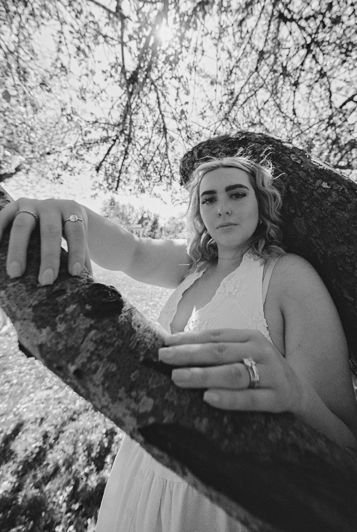 Black and white photo of a woman in white dress leaning on a tree branch wearing rings.