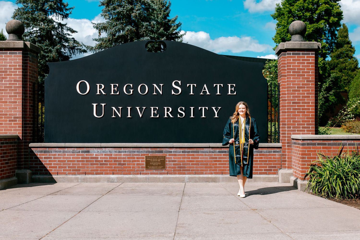 Graduate in cap and gown poses proudly in front of Oregon State University brick entrance sign on a sunny day.