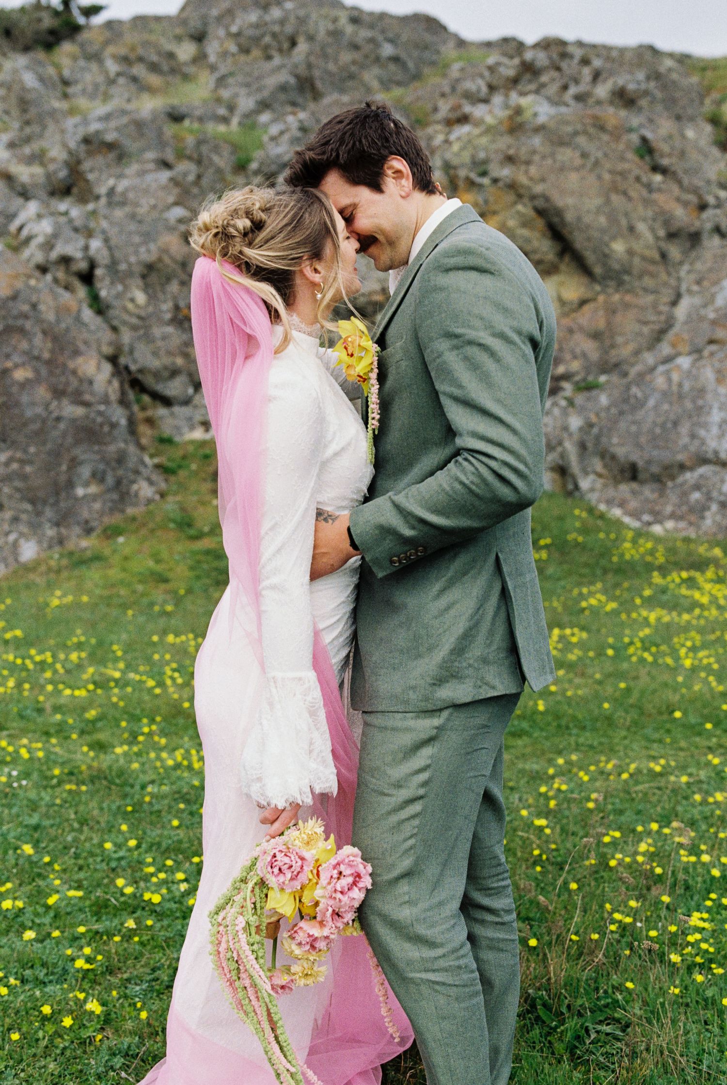 Bride in white gown with pink veil holds floral bouquet, touching foreheads with groom in green suit outdoors.