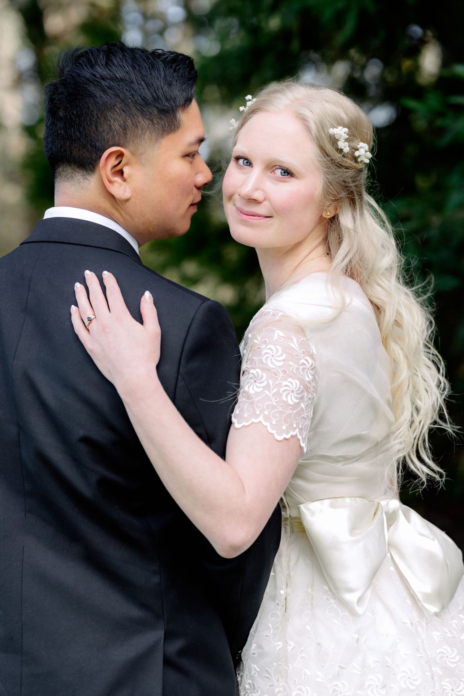 Blonde bride in lace wedding dress with large bow embraces groom in black suit outdoors.
