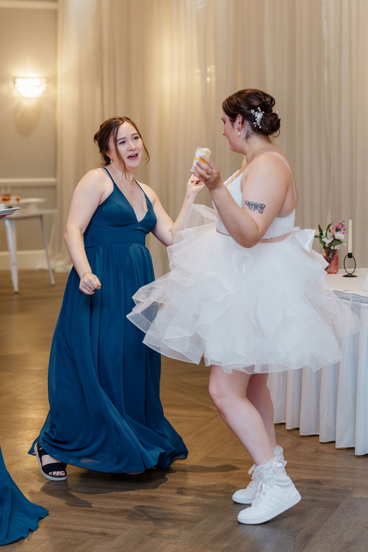 Bride in short white dress dancing energetically with bridesmaid in blue gown at wedding reception.