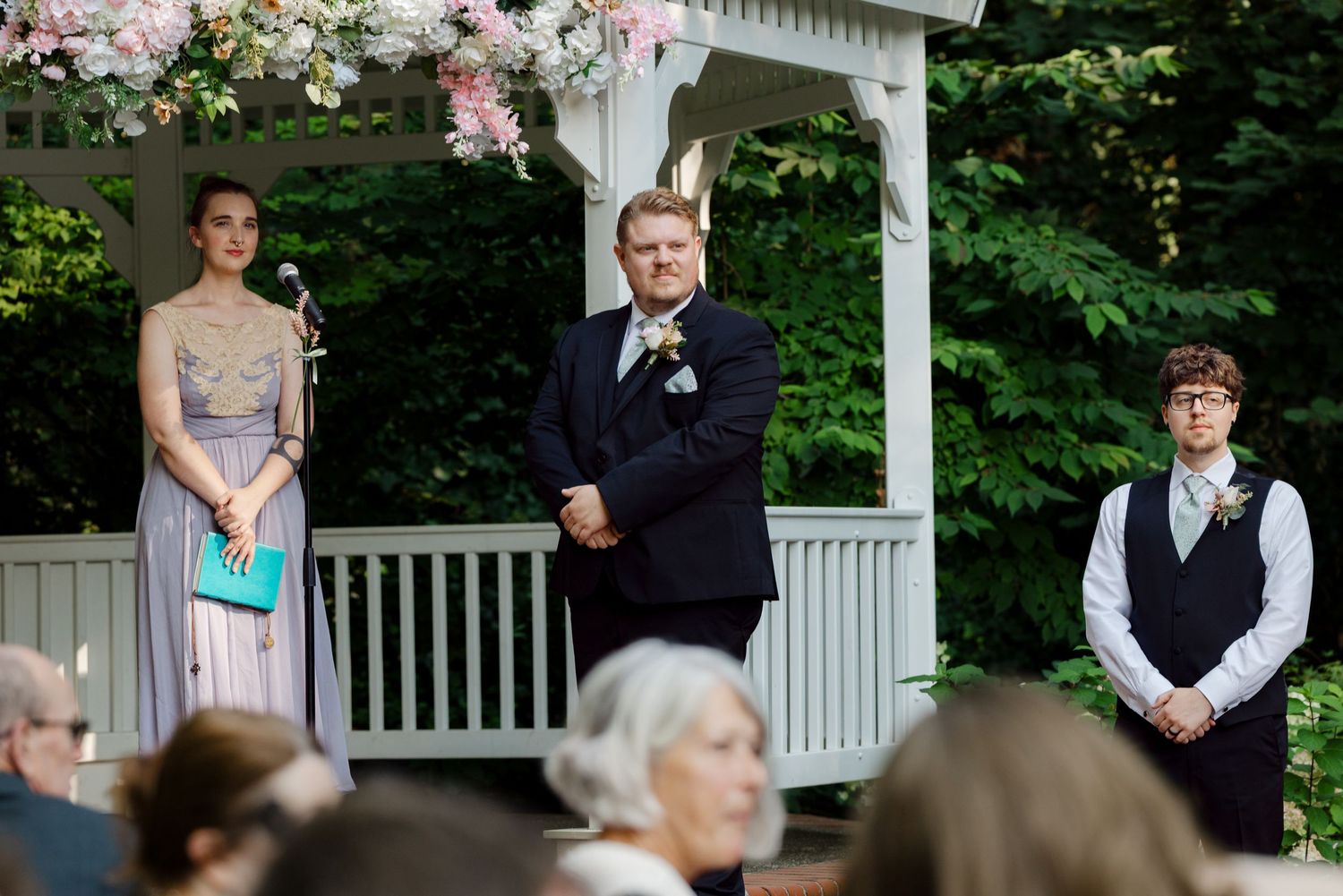 Wedding officiant and attendants standing at outdoor gazebo altar with floral decorations.