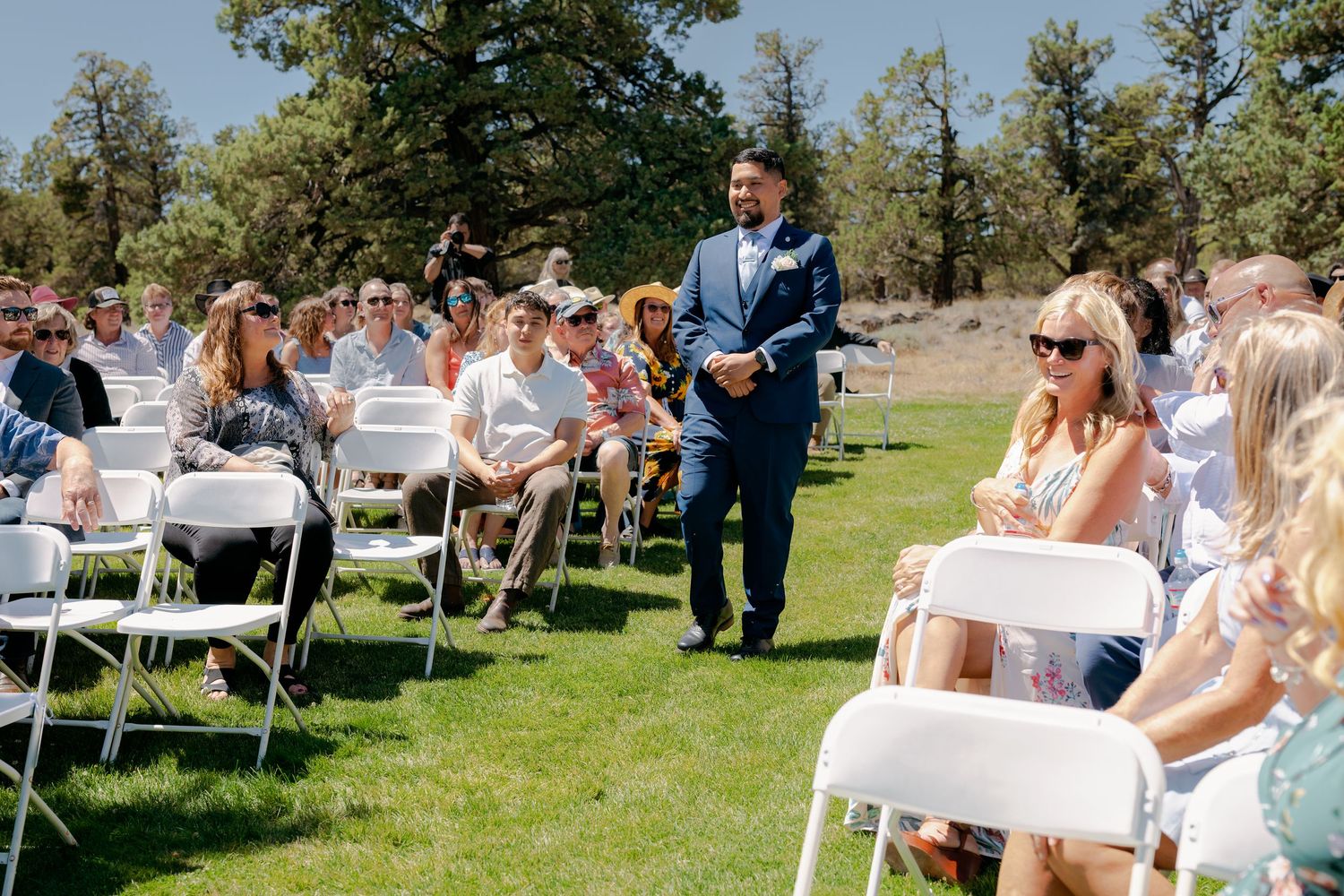 Groom in navy suit walks down outdoor wedding ceremony aisle between rows of white chairs.