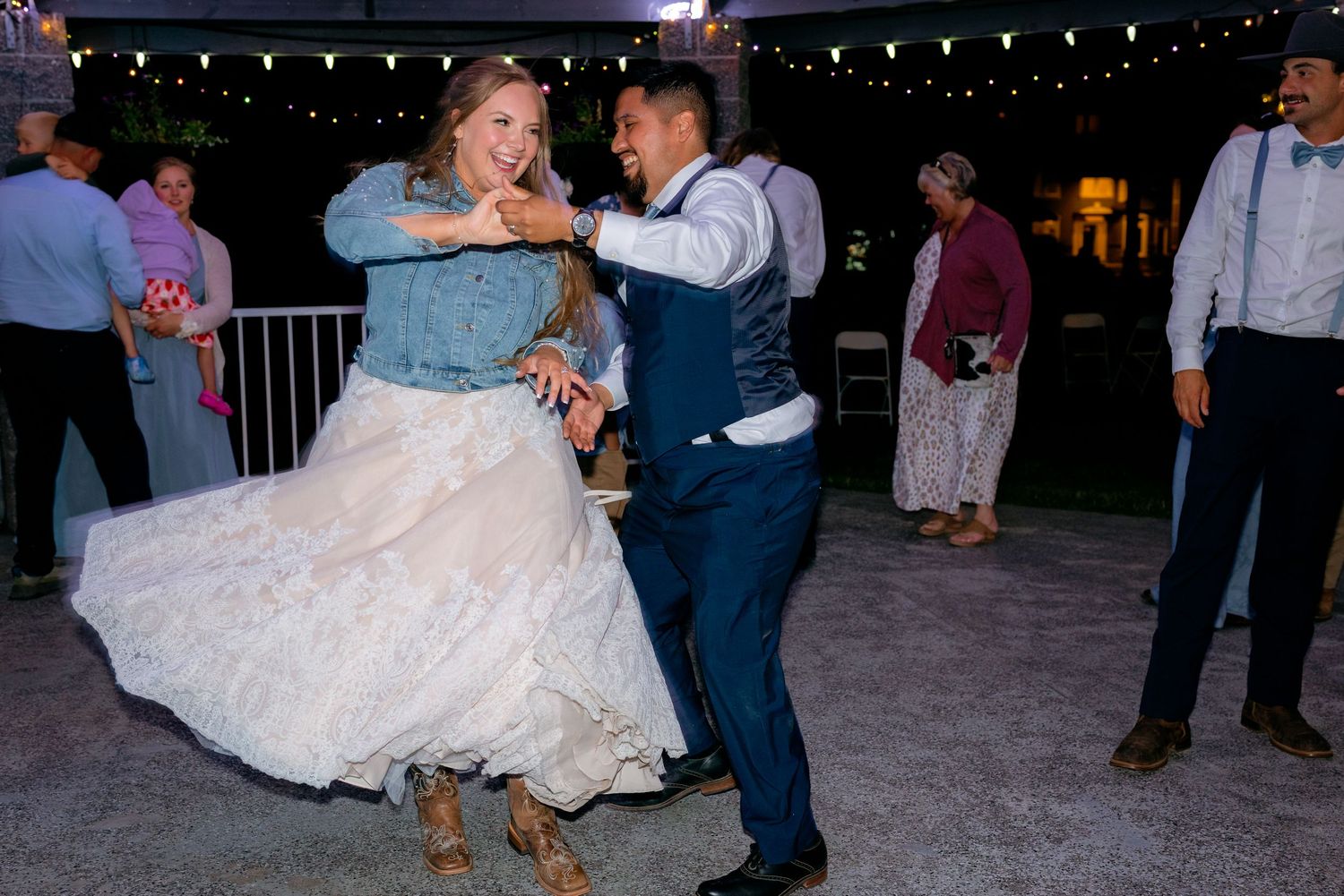 Bride in white dress and groom in navy vest dancing energetically at their outdoor wedding reception.