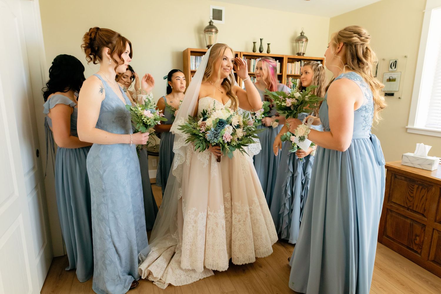 Bride in white gown surrounded by bridesmaids in dusty blue dresses holding floral bouquets during wedding preparations.