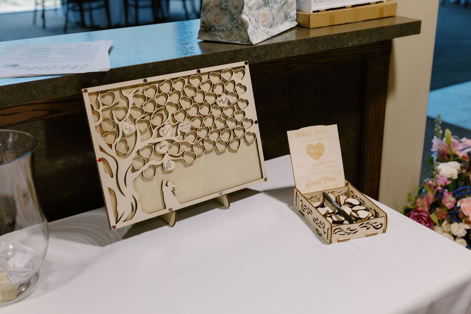 Wooden laser-cut wedding guest book tree with heart leaves displayed on table beside a box of wooden heart tokens.