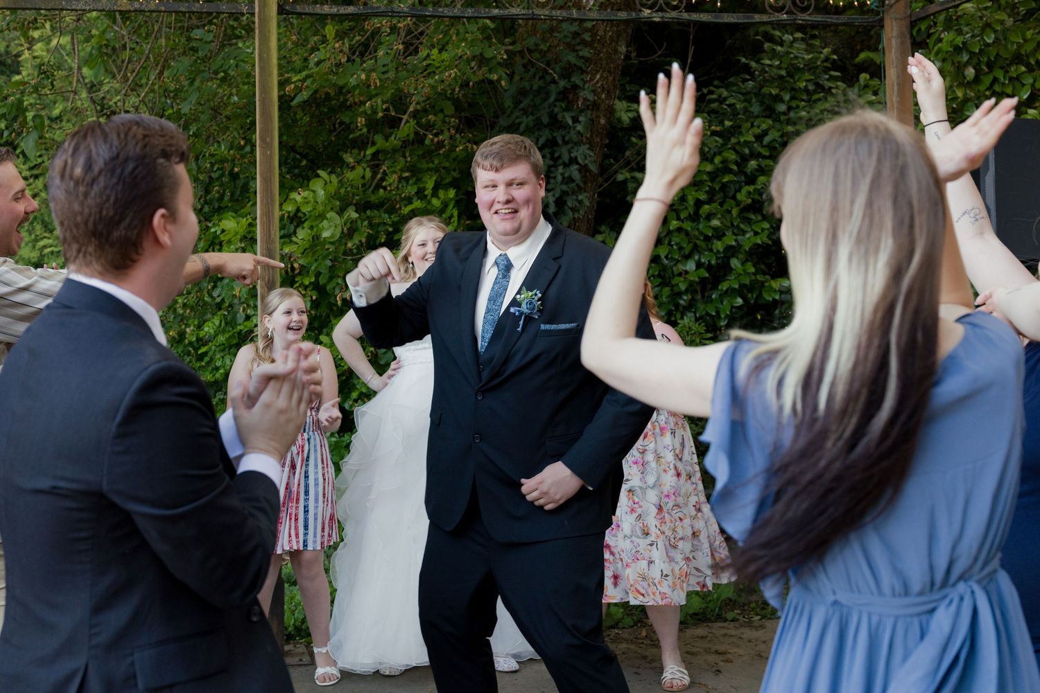 Groom celebrates joyfully with cheering wedding guests during lively outdoor reception dance.