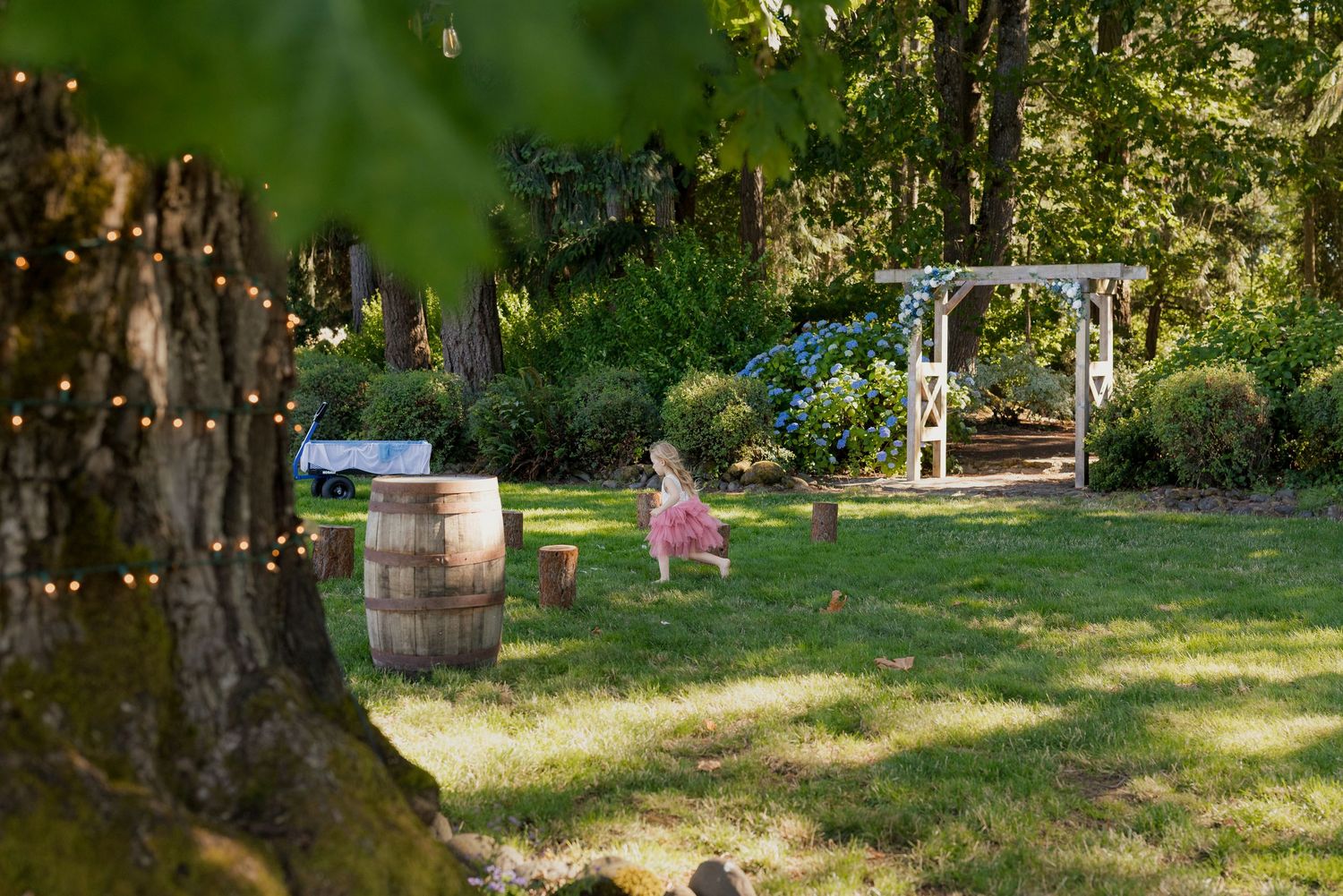 Child in pink dress plays in lush garden wedding venue with barrel and string lights.