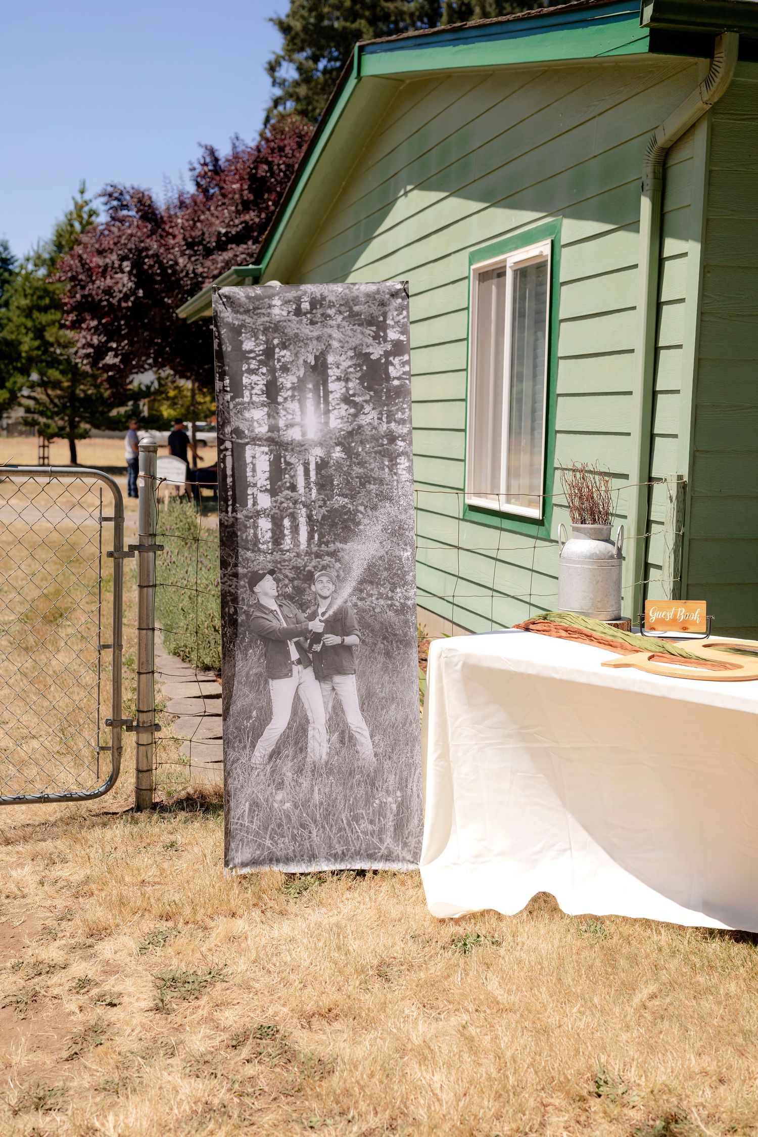 A black and white photo banner of two people stands beside a green farmhouse at an outdoor wedding reception.