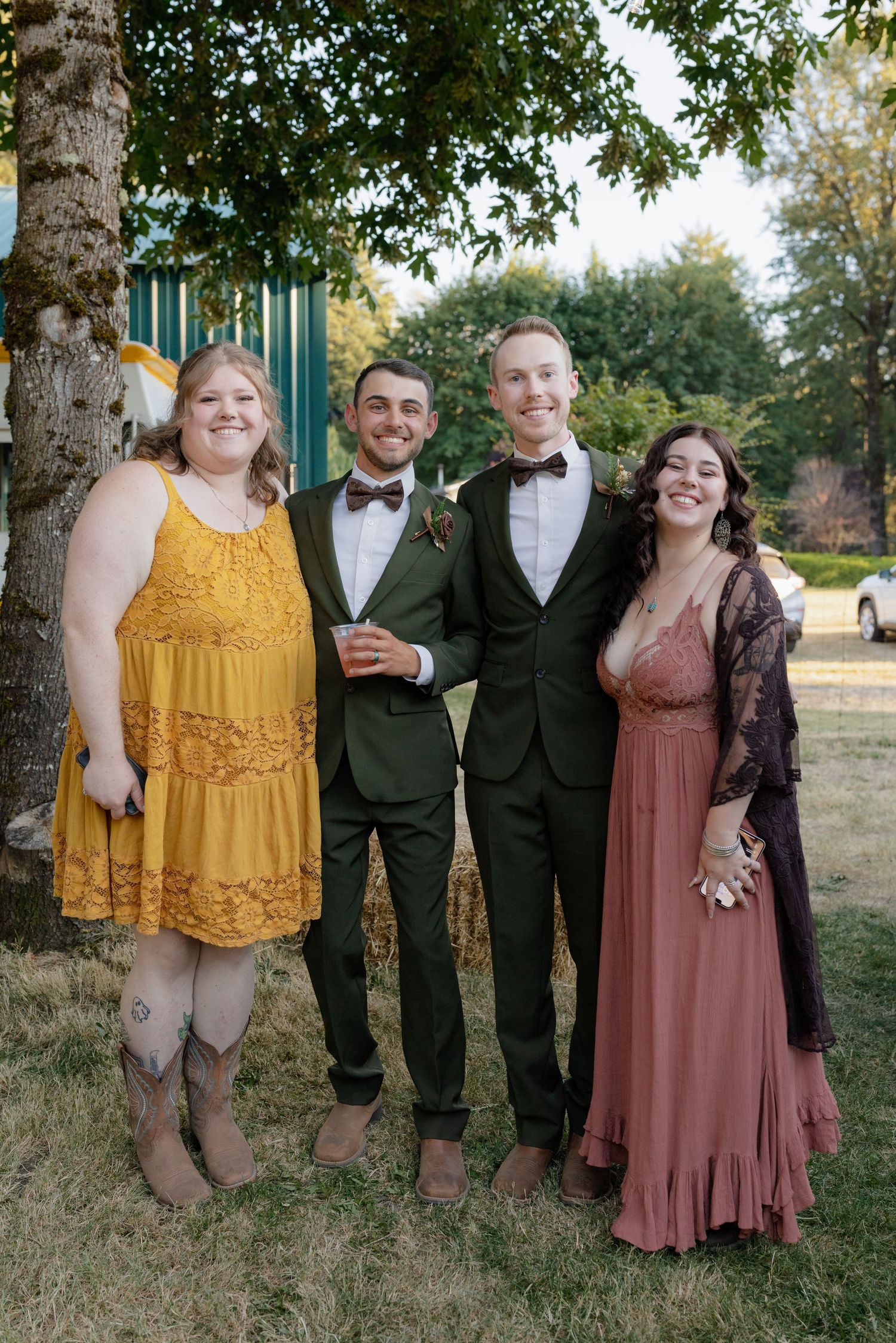 Two grooms in bow ties pose with two women in formal dresses outdoors at a wedding celebration.