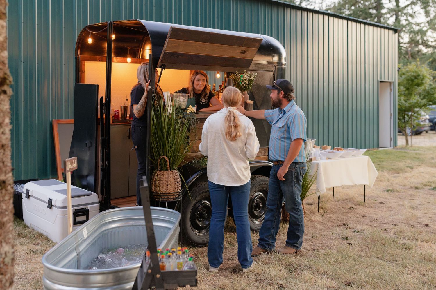 Guests gather around a vintage camper bar conversion at an outdoor rustic wedding reception venue.