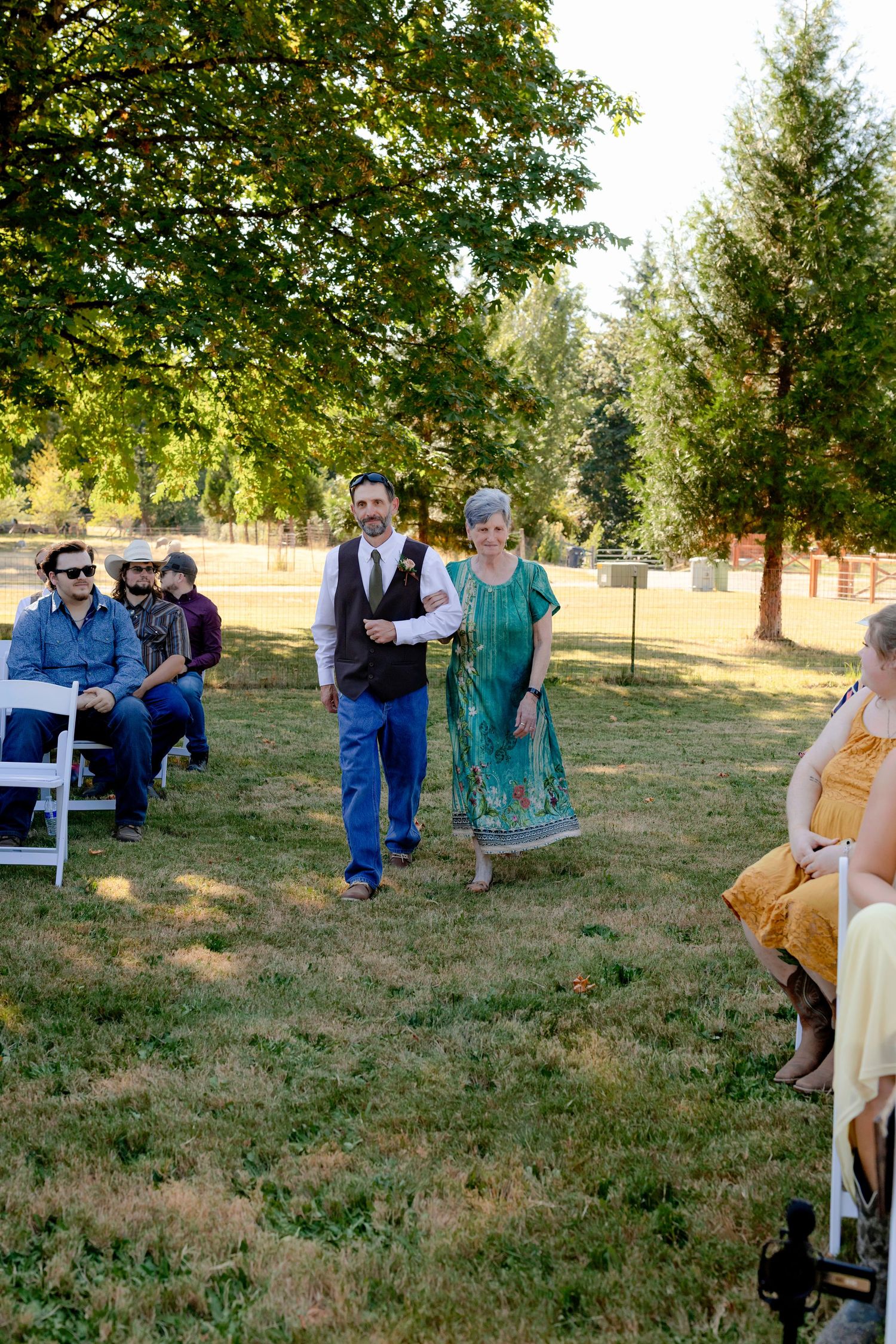 Groom escorted down outdoor wedding aisle by woman in teal dress among seated guests.
