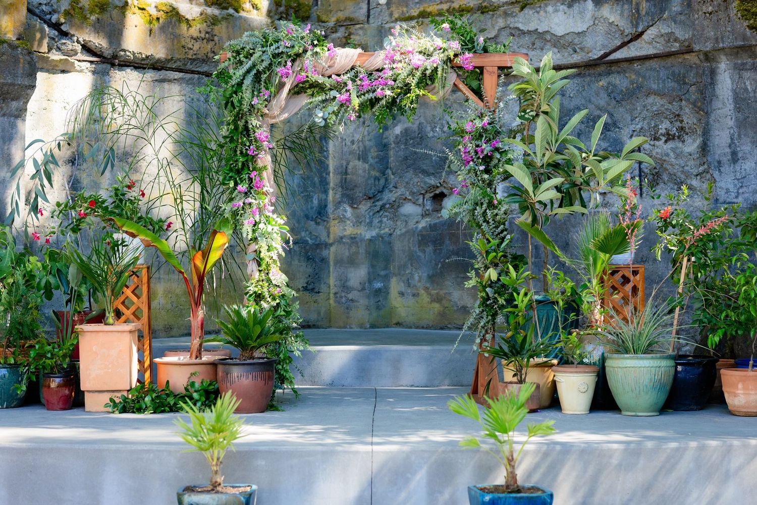 Lush garden display with potted tropical plants and a floral arch against a stone wall.