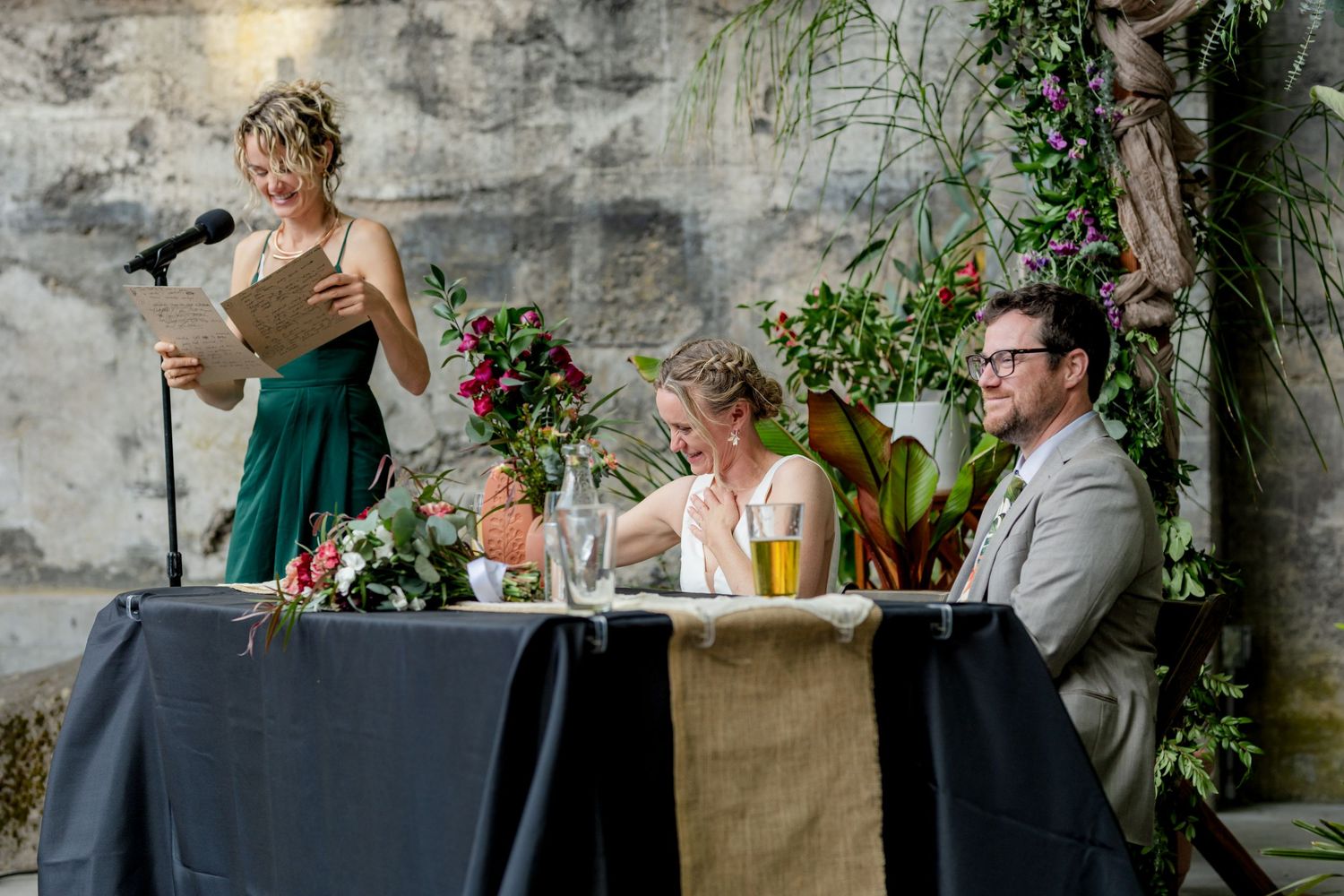 Bridesmaid in green dress gives speech at microphone while bride and groom laugh at sweetheart table.