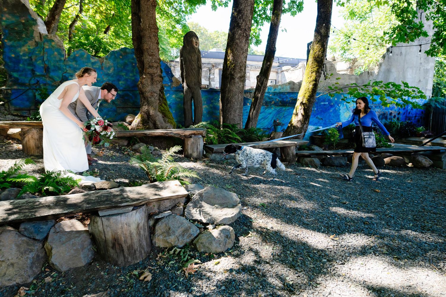 Bride and groom sawing wood together outdoors near stone walls and colorful mural, with guest walking nearby.