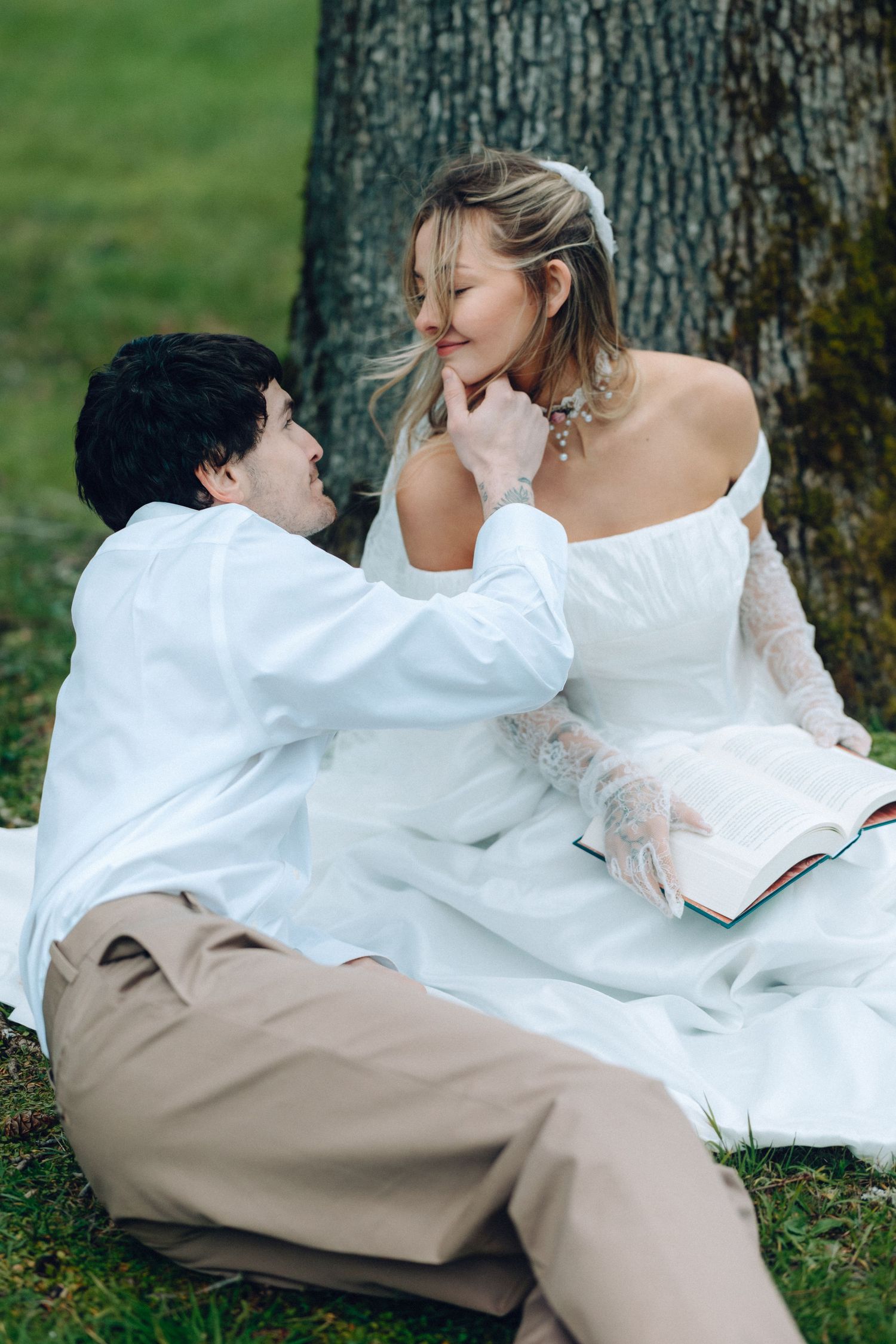 Wedding couple sharing intimate moments while sitting together on grass near a tree.