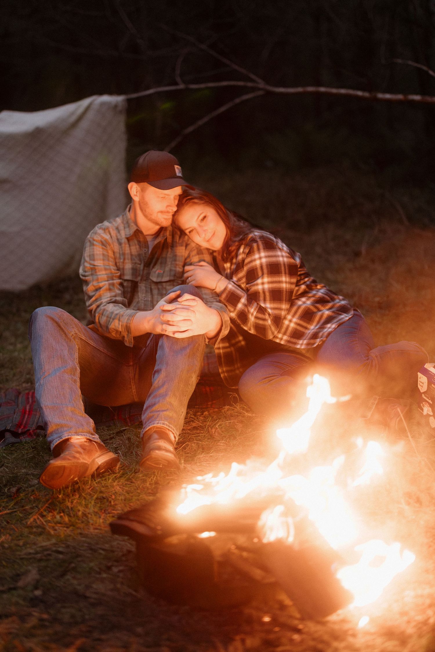 A couple sits close together by a warm campfire at night, wearing plaid shirts and enjoying the cozy outdoor atmosphere.