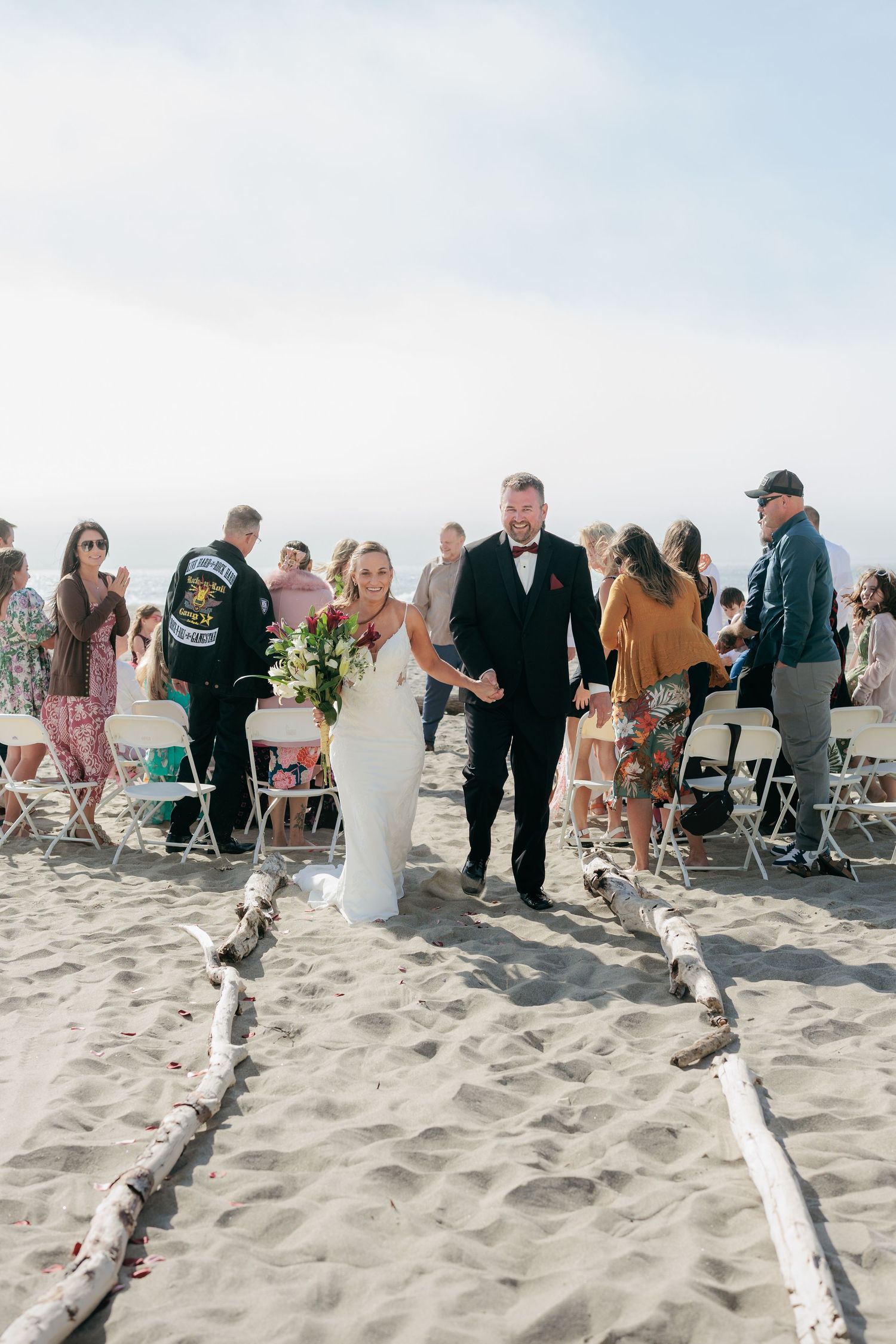 A beach wedding ceremony with guests seated on folding chairs and driftwood lining the sandy aisle.