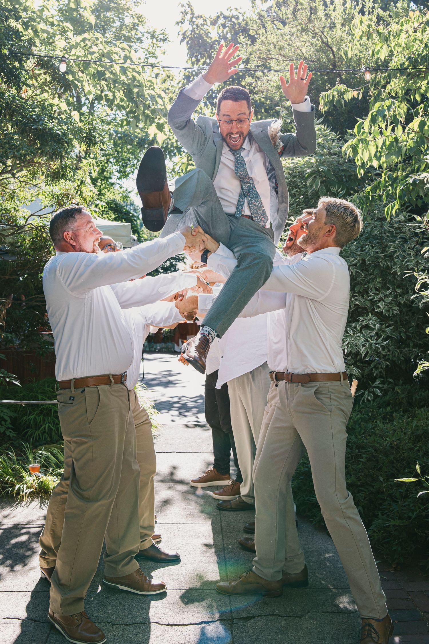 Groomsmen in khaki suits perform playful jumping poses for photos in a garden setting.