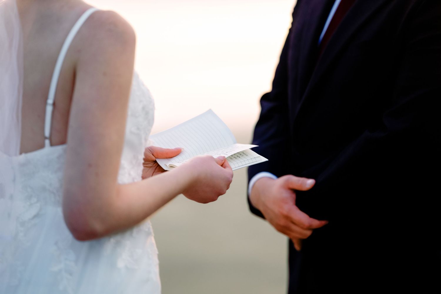 A bride in a white wedding dress holds a bouquet while standing in a sunlit room.