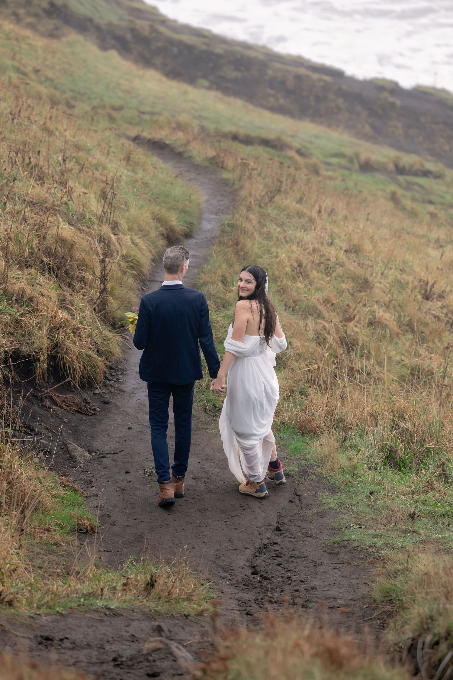 A couple in wedding attire walking hand in hand along a winding dirt path through grassy hillside on an overcast day.