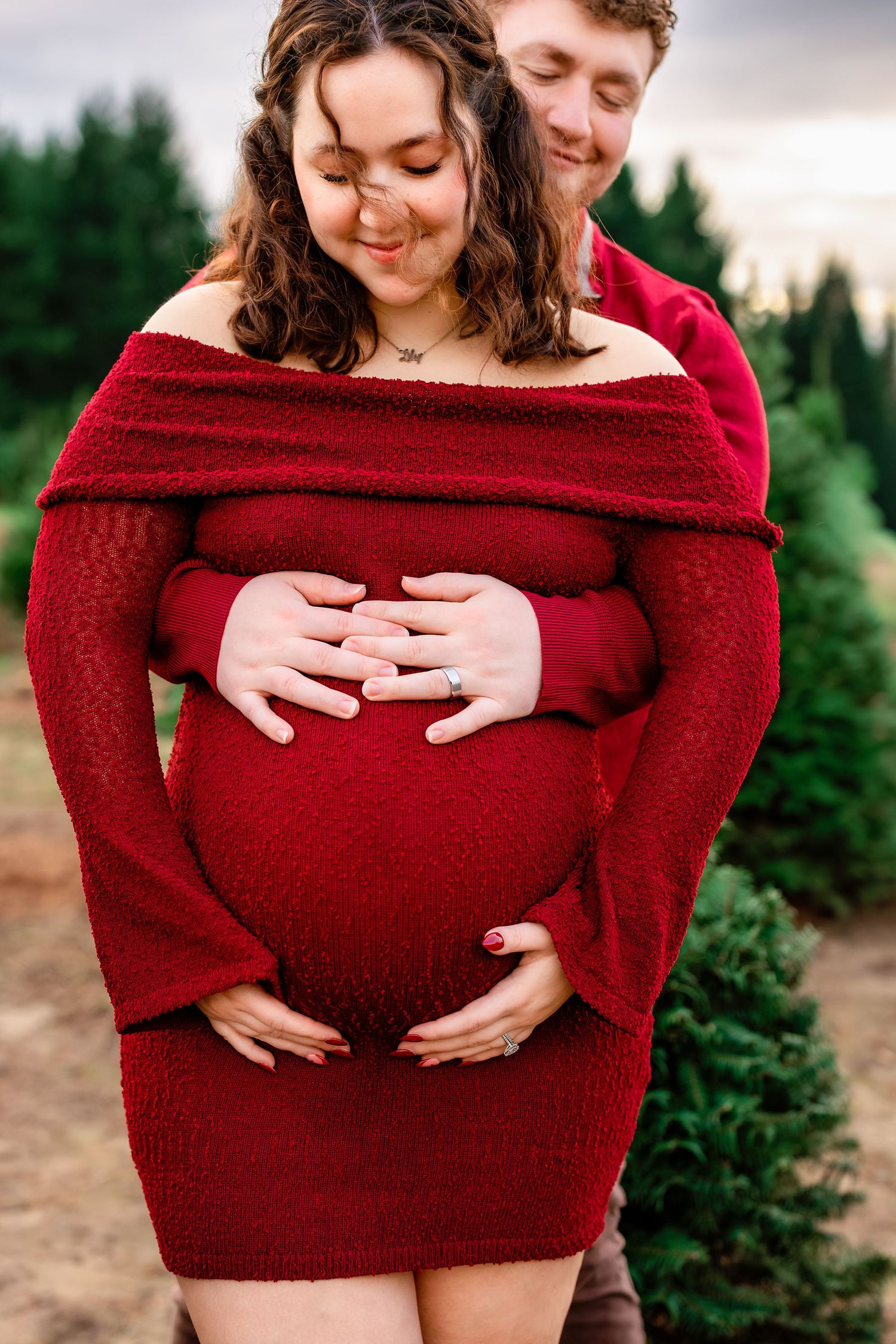 A couple shares an intimate maternity photo session among evergreen trees wearing a red off-shoulder sweater.