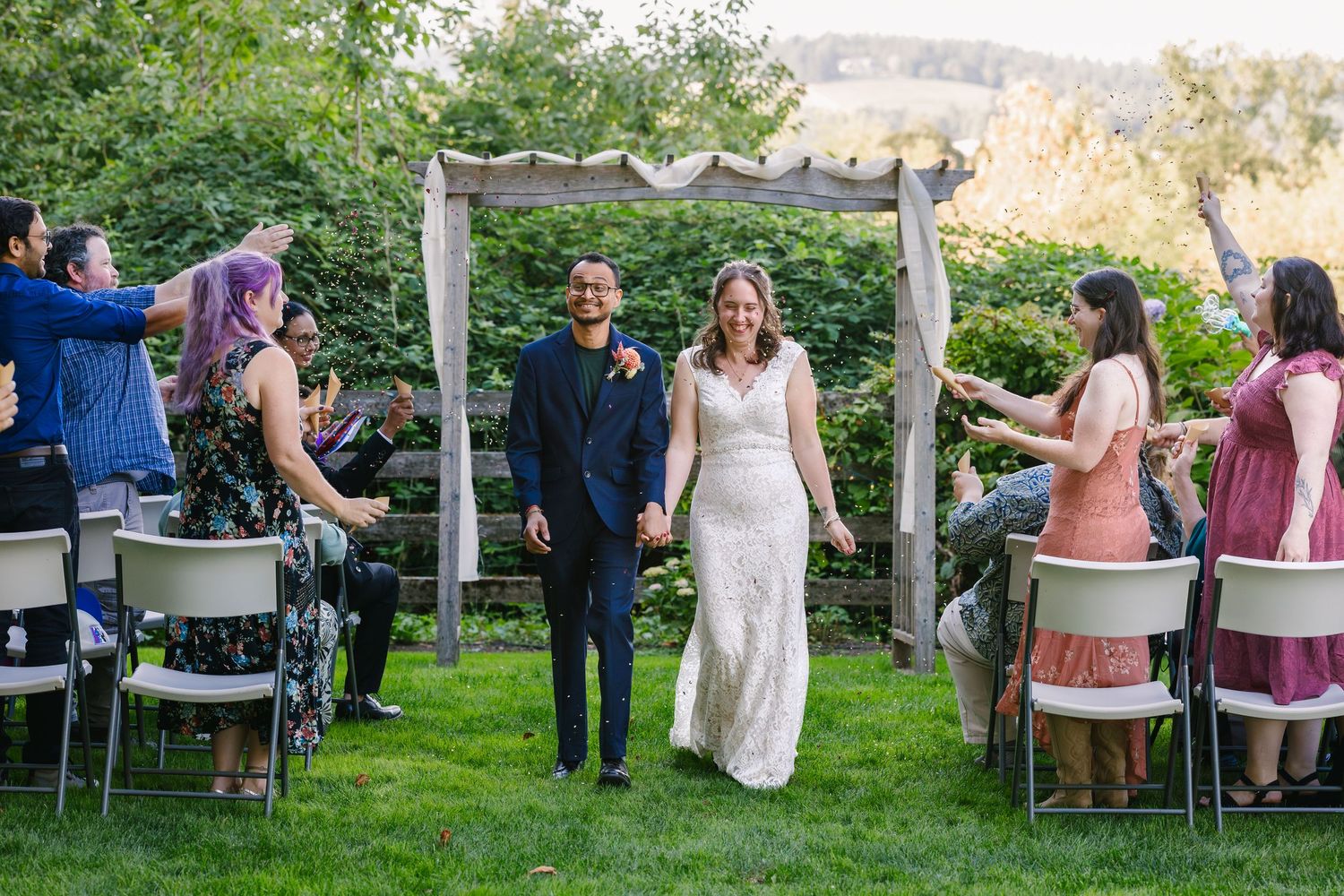 Outdoor garden party setup with white chairs and tables arranged on a lawn surrounded by trees.