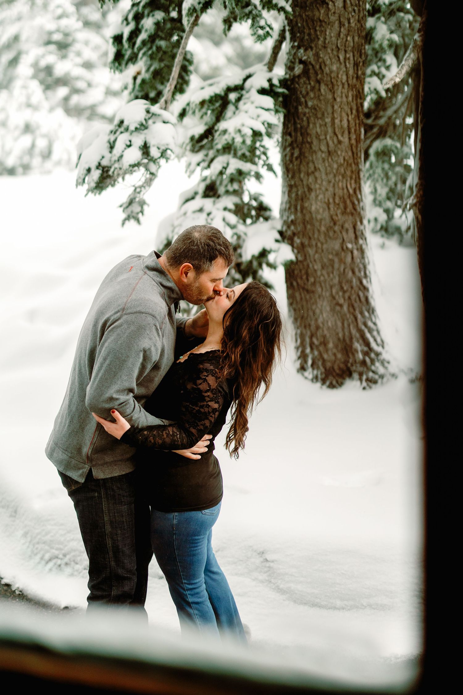 A romantic winter photoshoot shows a couple embracing among snow-covered evergreen trees.