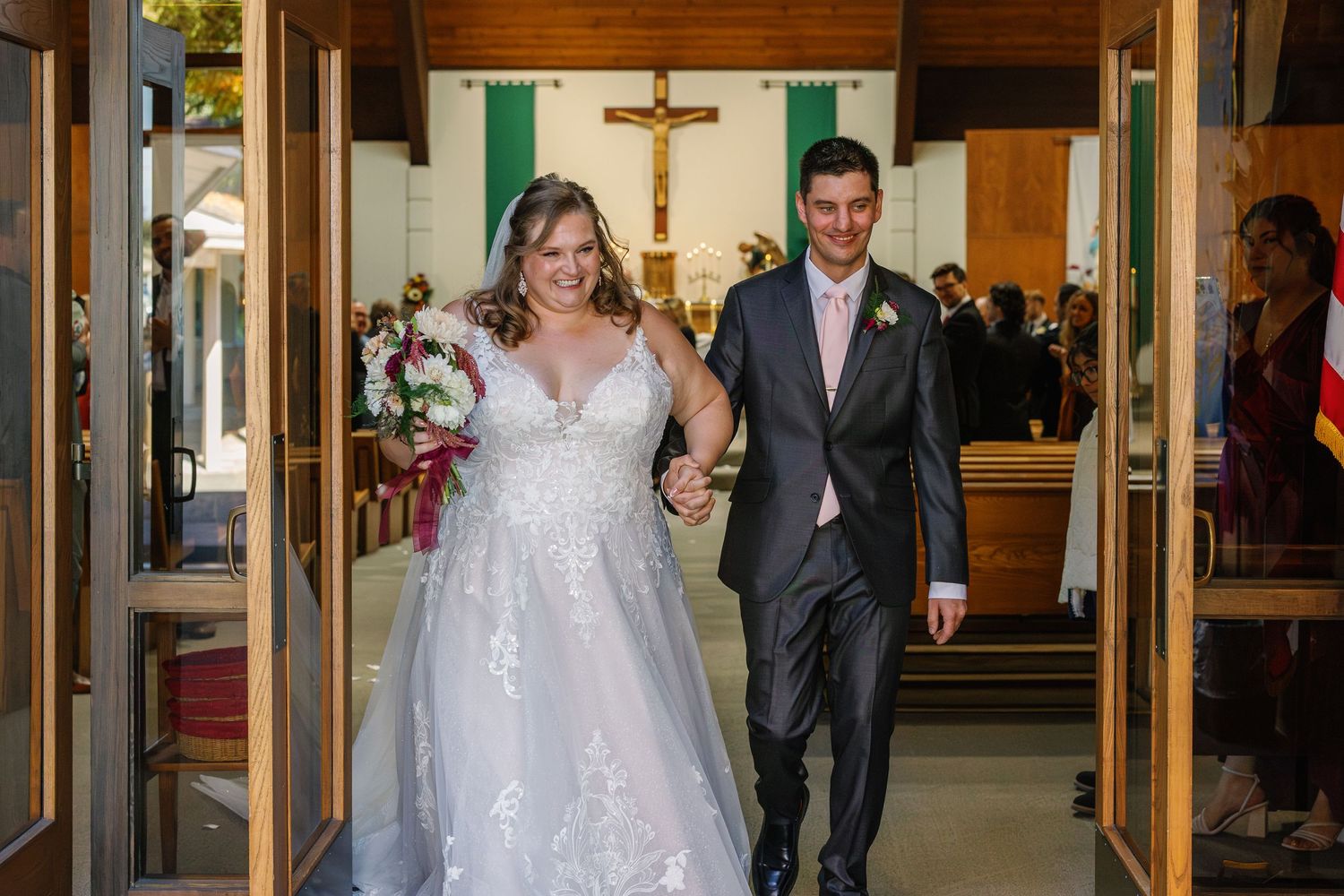 Wedding couple walks down church hallway with wooden paneled walls and glass doors.