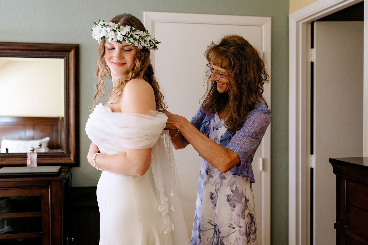 Bride in white gown and floral crown gets veil adjusted by smiling woman in blue floral dress before wedding.