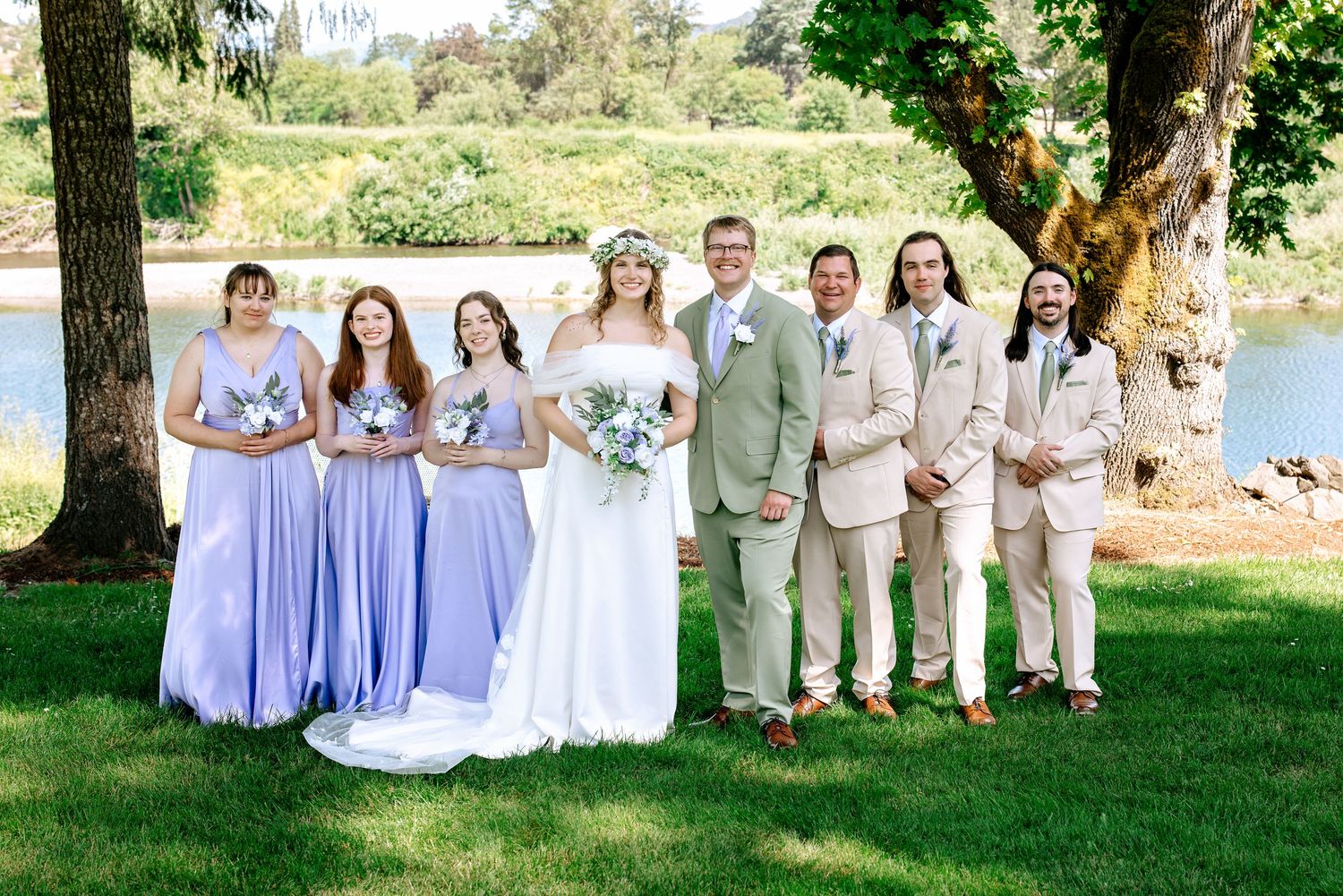 Wedding party posing by lake with bridesmaids in lavender gowns and groomsmen in tan suits.