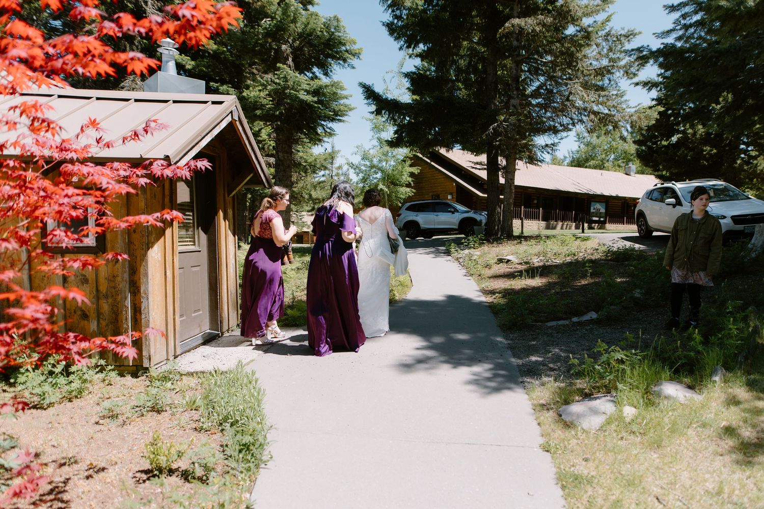 Bride in white gown with bridesmaids in purple dresses gathered outside rustic wedding venue on sunny day.