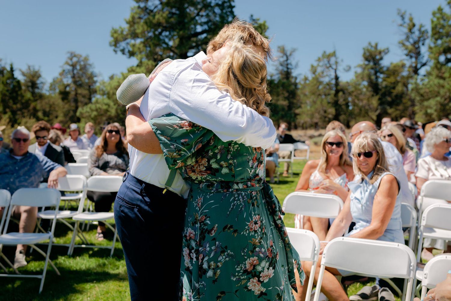 Groom hugging woman in green floral dress at outdoor wedding ceremony with white chairs.