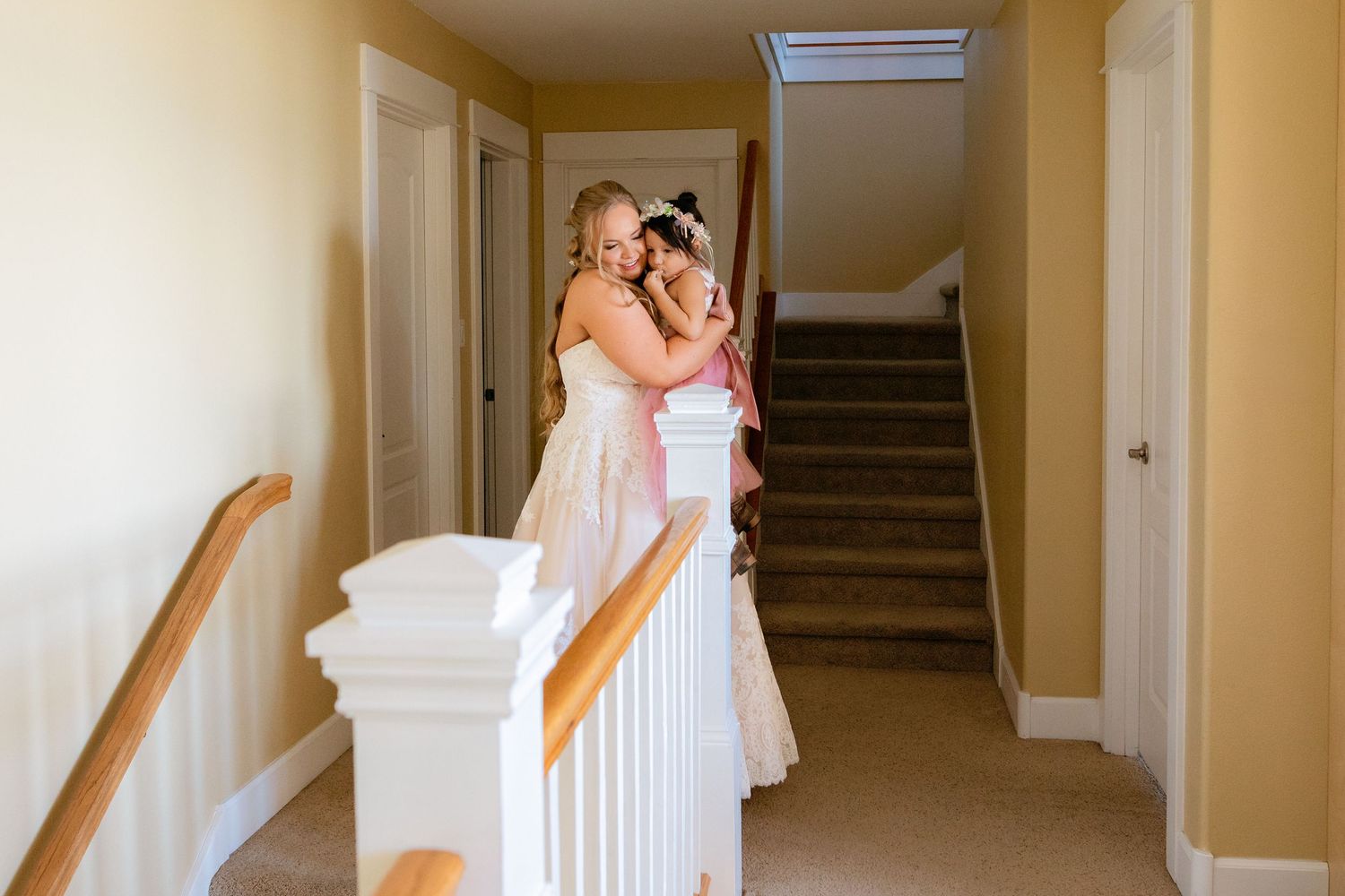 Two young girls in pink dresses sharing a sweet hug on a staircase landing in a bright home.