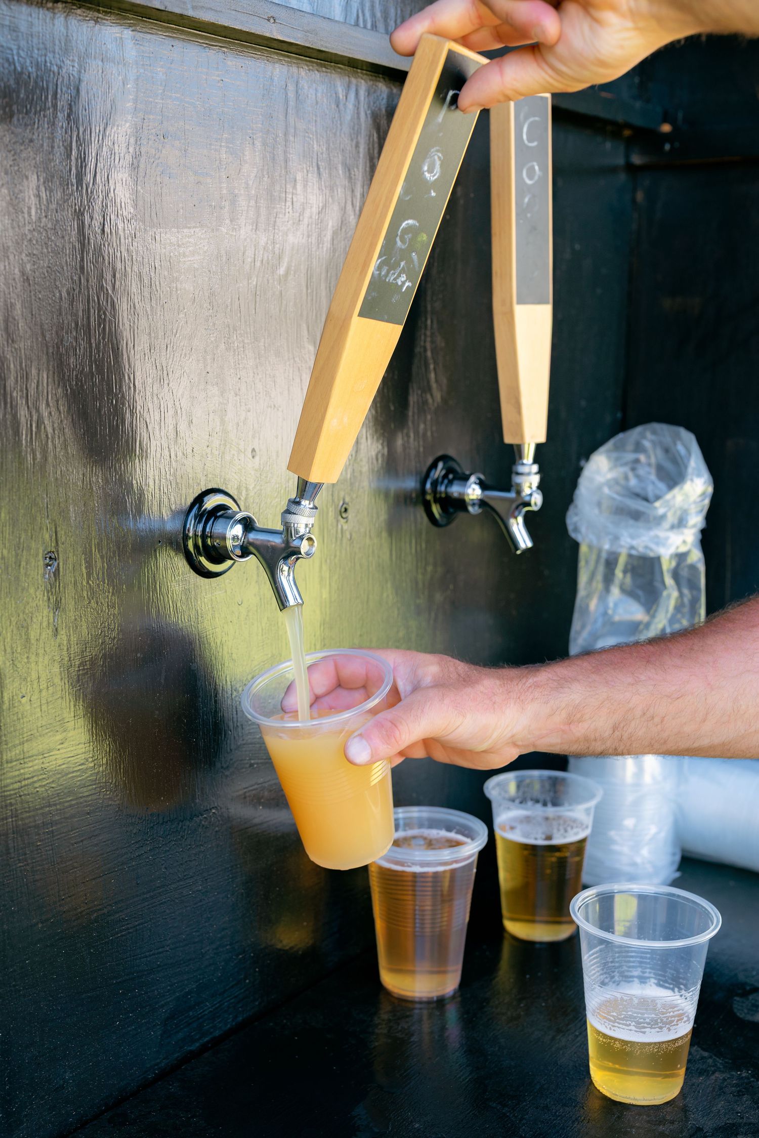 Bartender pouring golden beer from a tap into plastic cups at an outdoor wedding reception bar.
