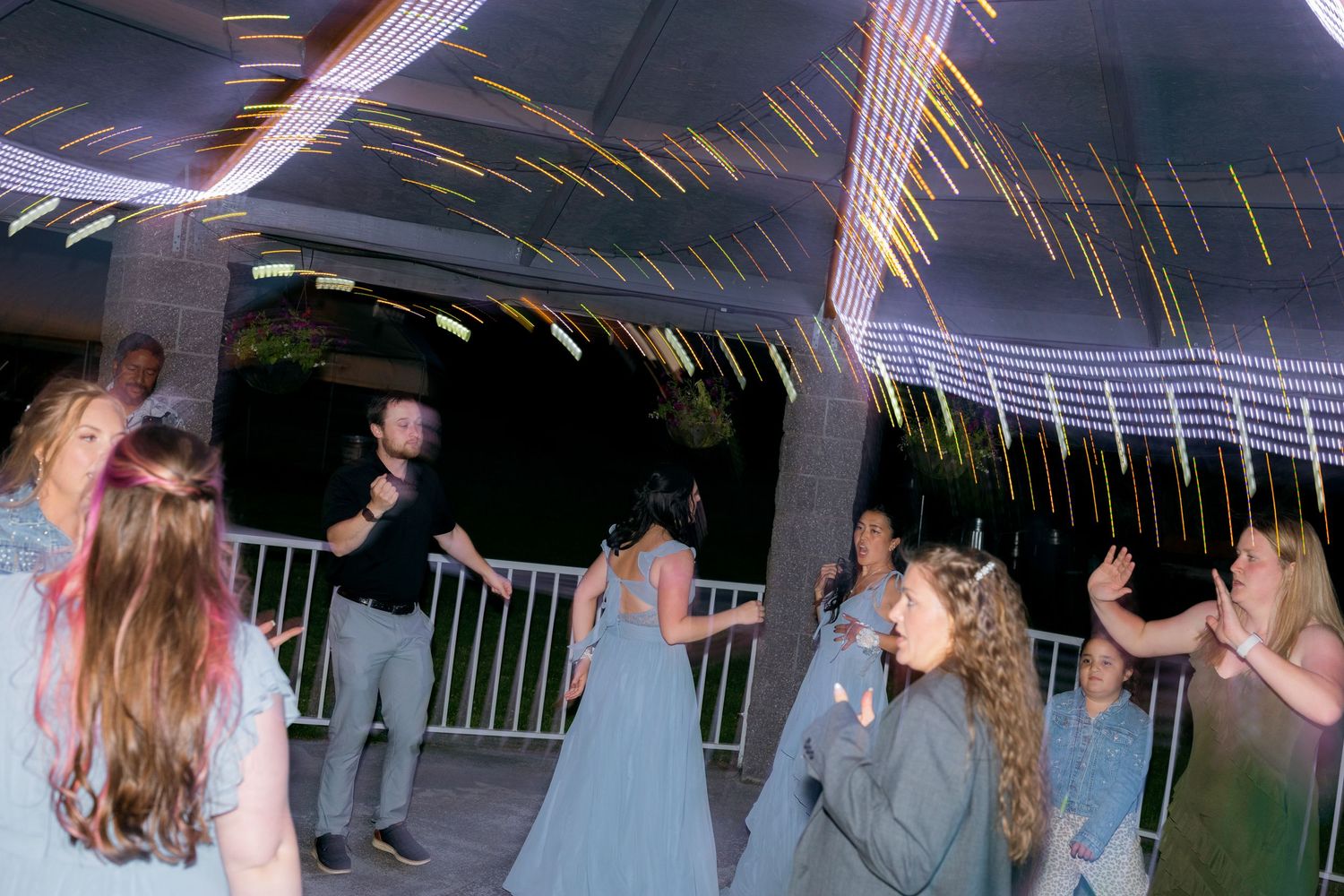 Wedding guests dancing under colorful light streaks at an outdoor evening reception party.