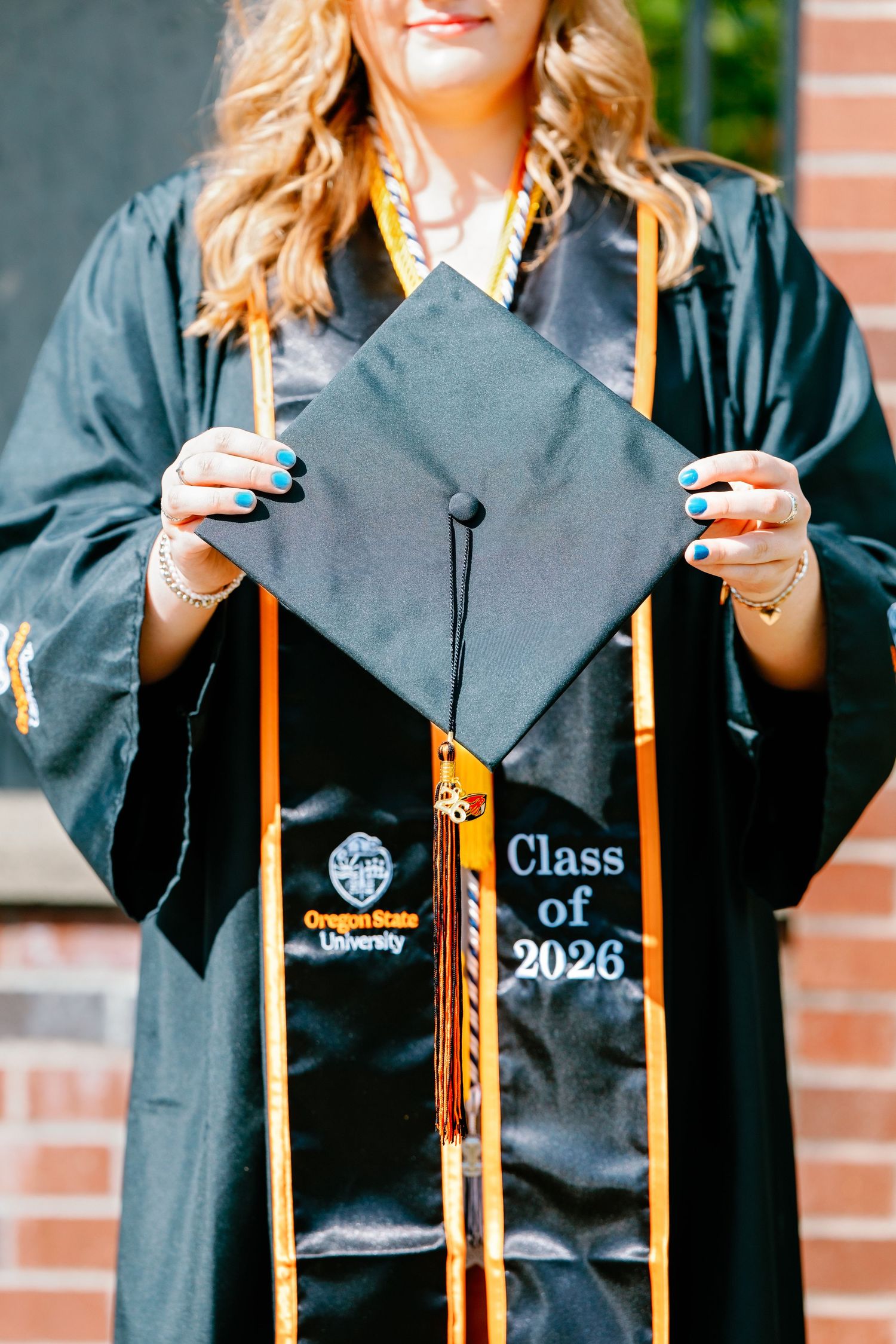 Oregon State University graduate holds black mortarboard cap displaying Class of 2026 while wearing gold honor stole.